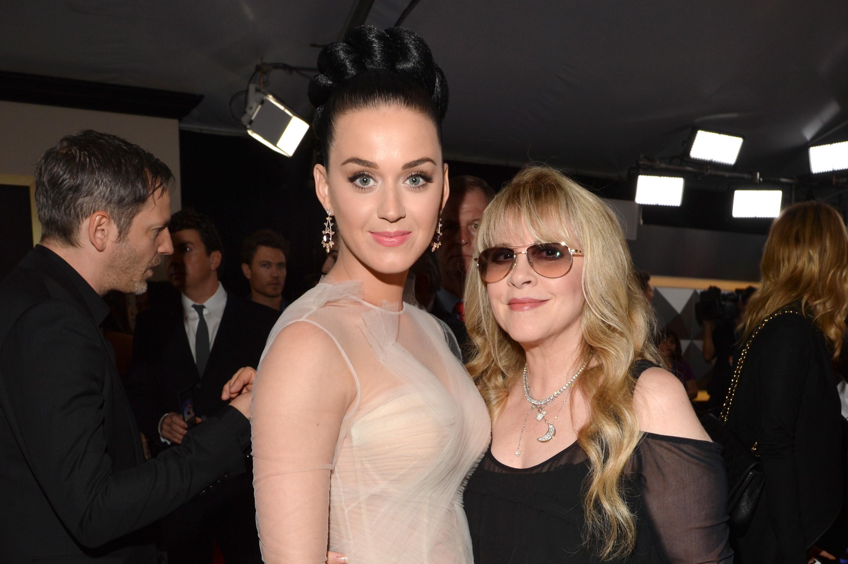 Katy Perry wears a white dress and stands next to Stevie Nicks who wears a black dress and sunglasses.