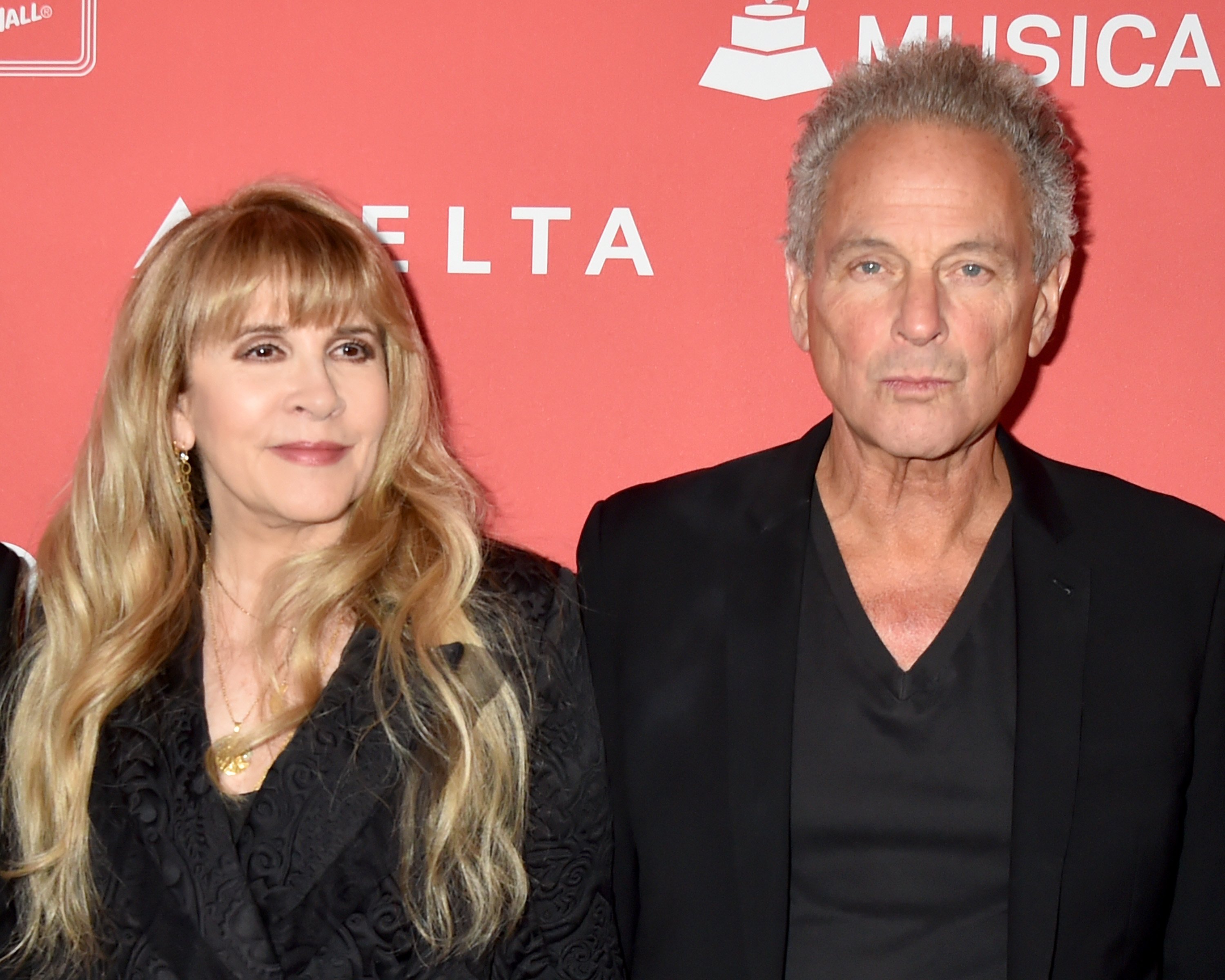 Stevie Nicks and Lindsey Buckingham both wear black and stand in front of a red background. 
