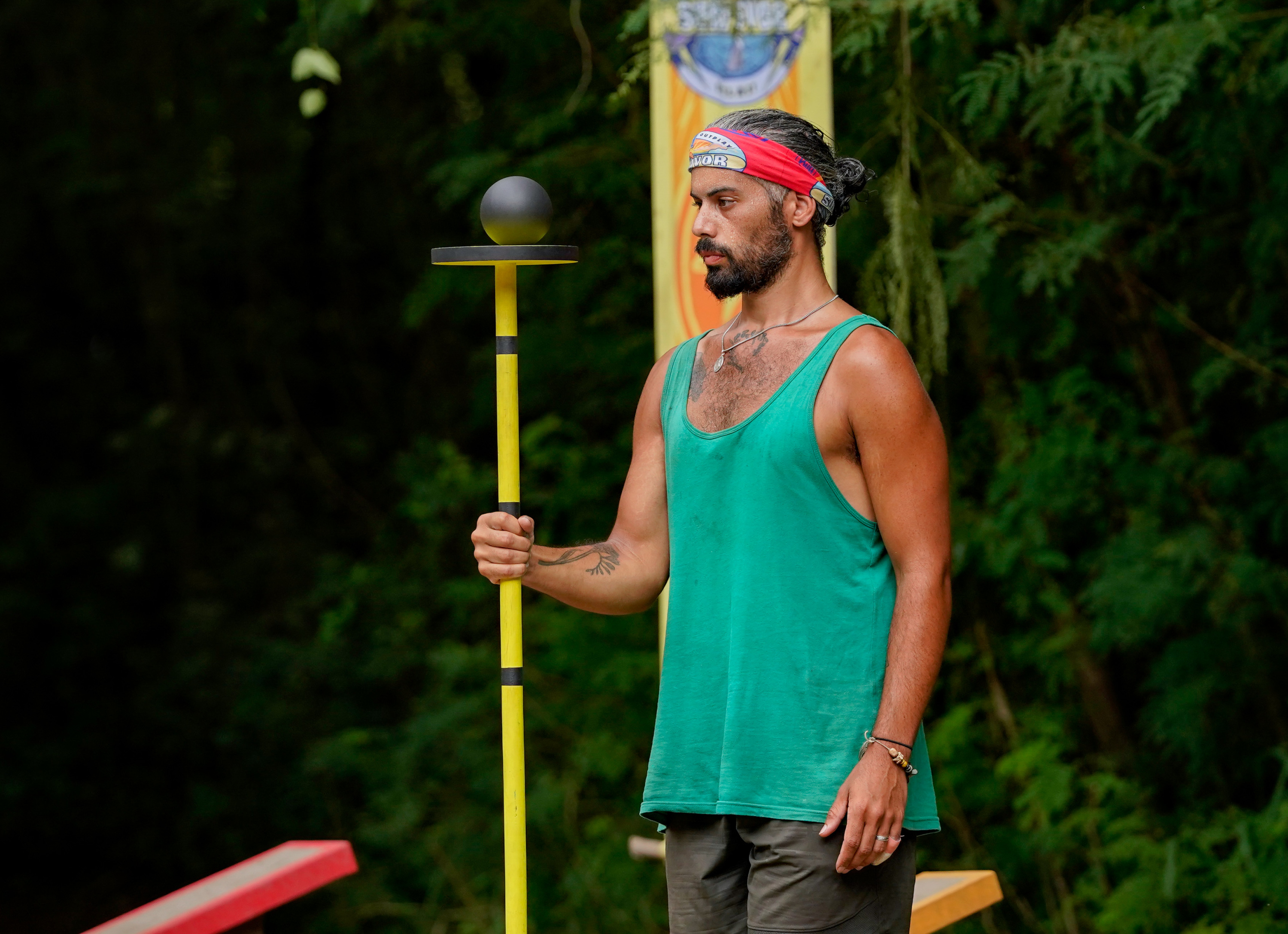 'Survivor' Season 41 star Ricard Foyé, who knows a cast member from season 42, wears a green tank top, gray shorts, and a red 'Survivor' bluff on his head while trying to balance a ball on a stick for a challenge.