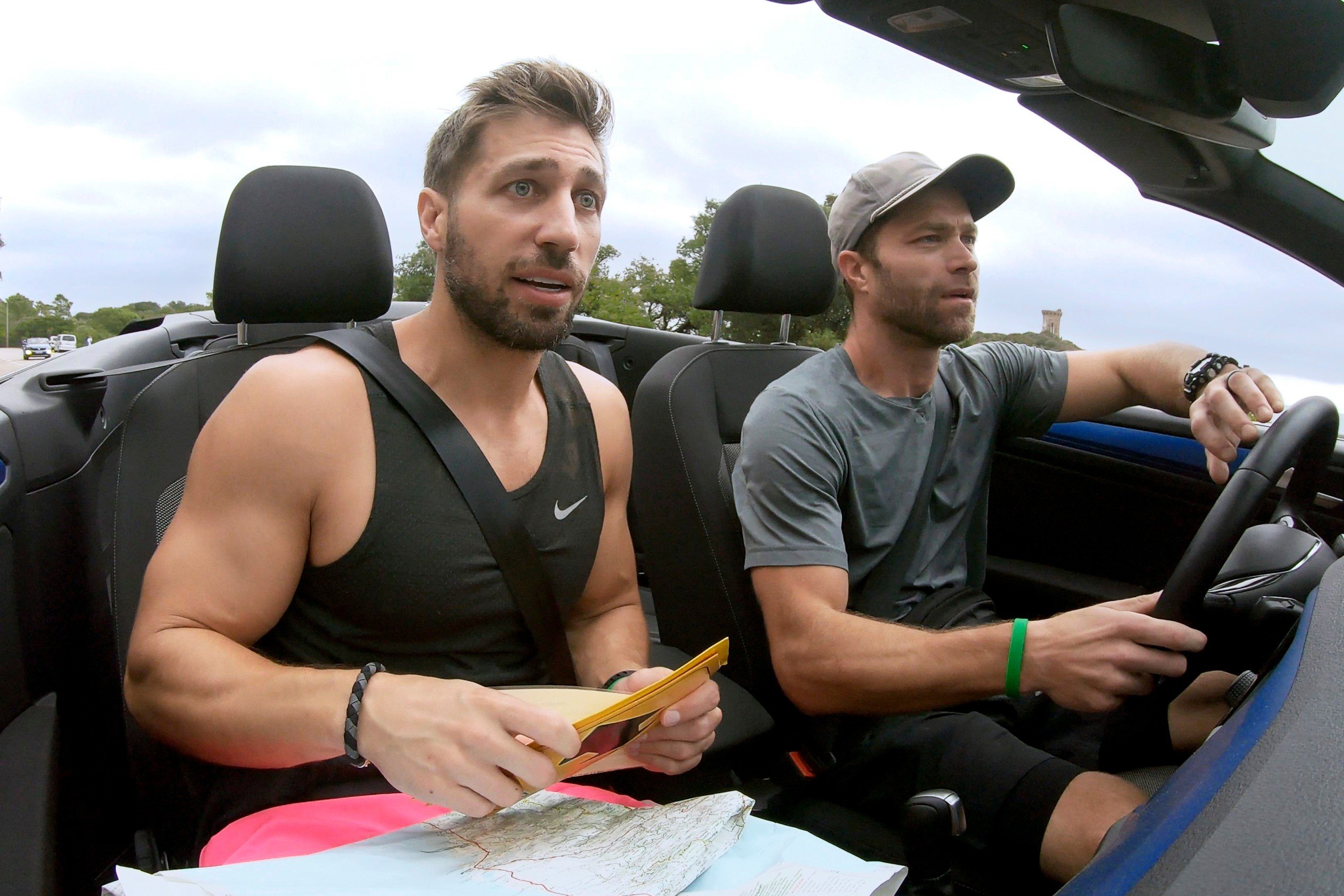 'The Amazing Race' Season 33 duo Ryan Ferguson and Dusty Harris ride in a car. Ryan wears a black tank top and pink shorts. Dusty wears a gray shirt, black shorts, and a gray hat.