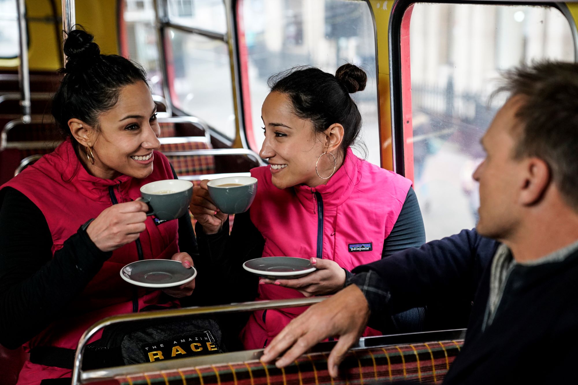 'The Amazing Race' Season 33 stars Lulu and Lala Gonzalez talk with host Phil Keoghan at a pit stop. Lulu and Lala wear pink vests over black long-sleeved shirts, and they sip cups of tea. Keoghan wears a black  coat.