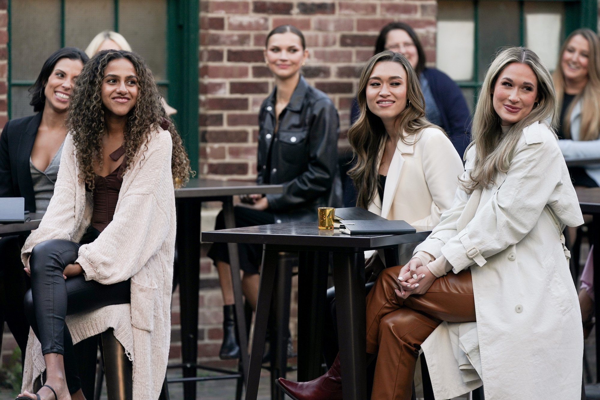 The women of 'The Bachelor' Season 26 sit at tables outside in front of a brick wall.