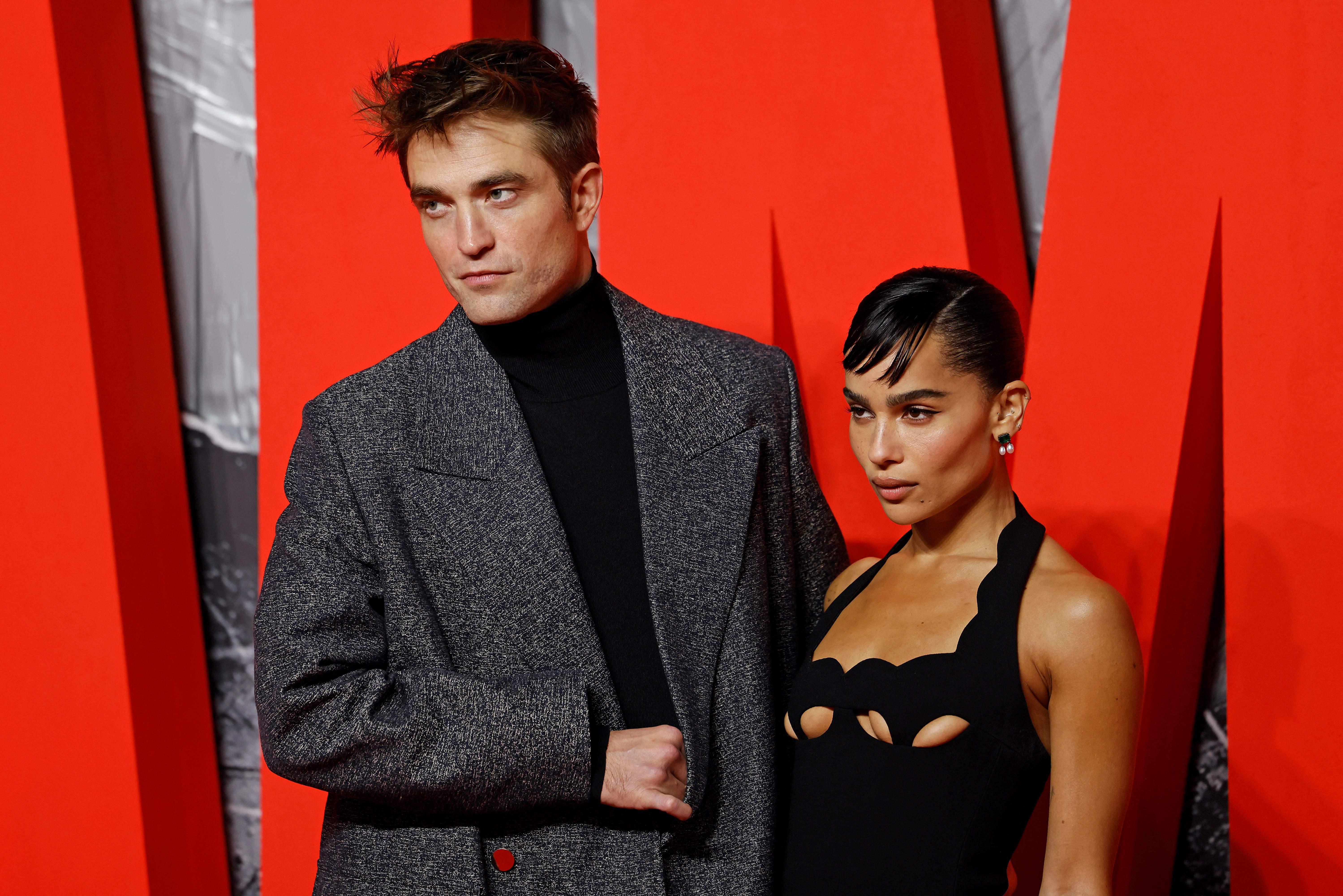 Robert Pattinson and Zoë Kravitz pose for photos together on the red carpet for 'The Batman' premiere. Pattinson wears a large gray coat over a black turtleneck. Kravitz wears a black dress.