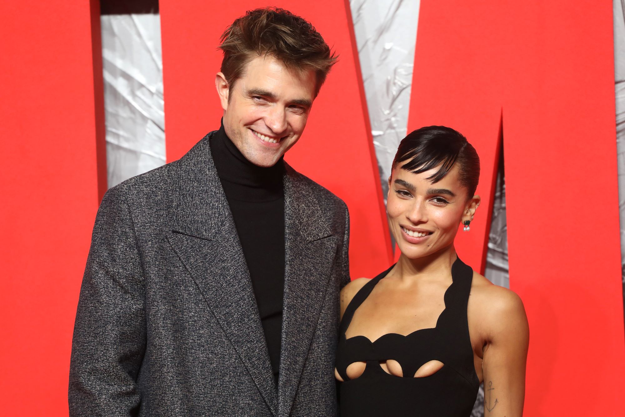 'The Batman' stars Robert Pattinson and Zoe Kravitz pose for pictures on a red carpet. Pattinson wears a gray coat over a black turtleneck. Kravitz wears a black dress.