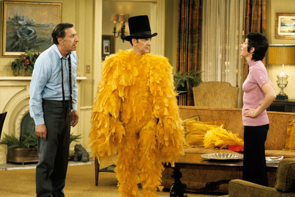 Actor Tony Randall, center, wears a top hat and feathery full-body yellow costume in a scene from 'The Odd Couple.' Actors Jack Klugman, left, and Elinor Donahue, right, appear with him.