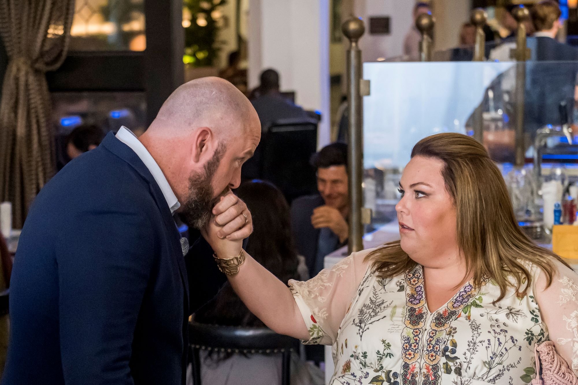 'This Is Us' Season 6 stars Chris Sullivan and Chrissy Metz, in character as Toby and Kate, share a scene where Toby kisses Kate's hand. Toby wears a blue suit. Kate wears a white dress with multi-colored floral patterns on it.