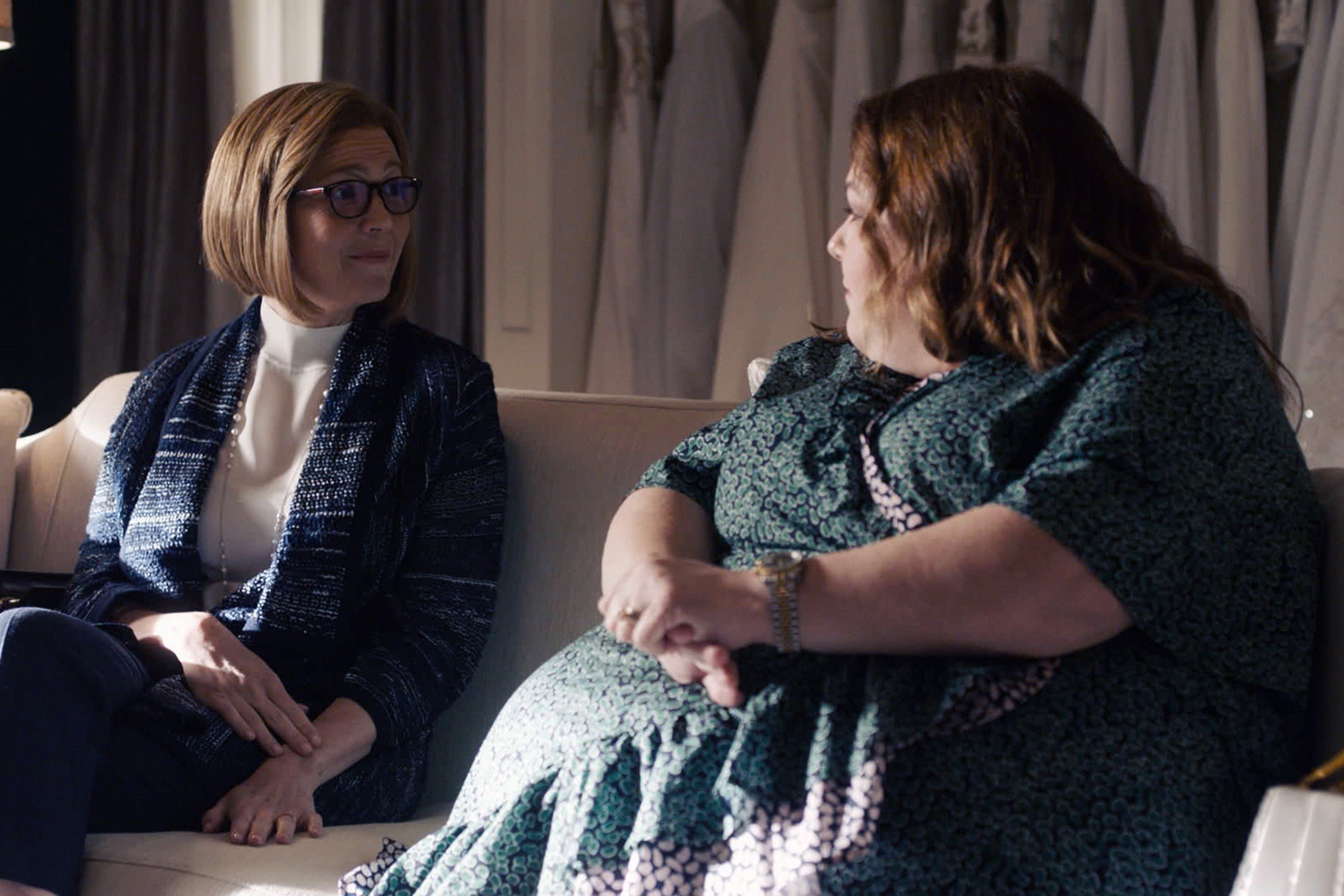 'This Is Us' stars Mandy Moore and Chrissy Metz, in character as Rebecca and Kate, share a scene. Rebecca wears a dark blue cardigan over a white turtleneck and dark pants. Kate wears a green floral dress.
