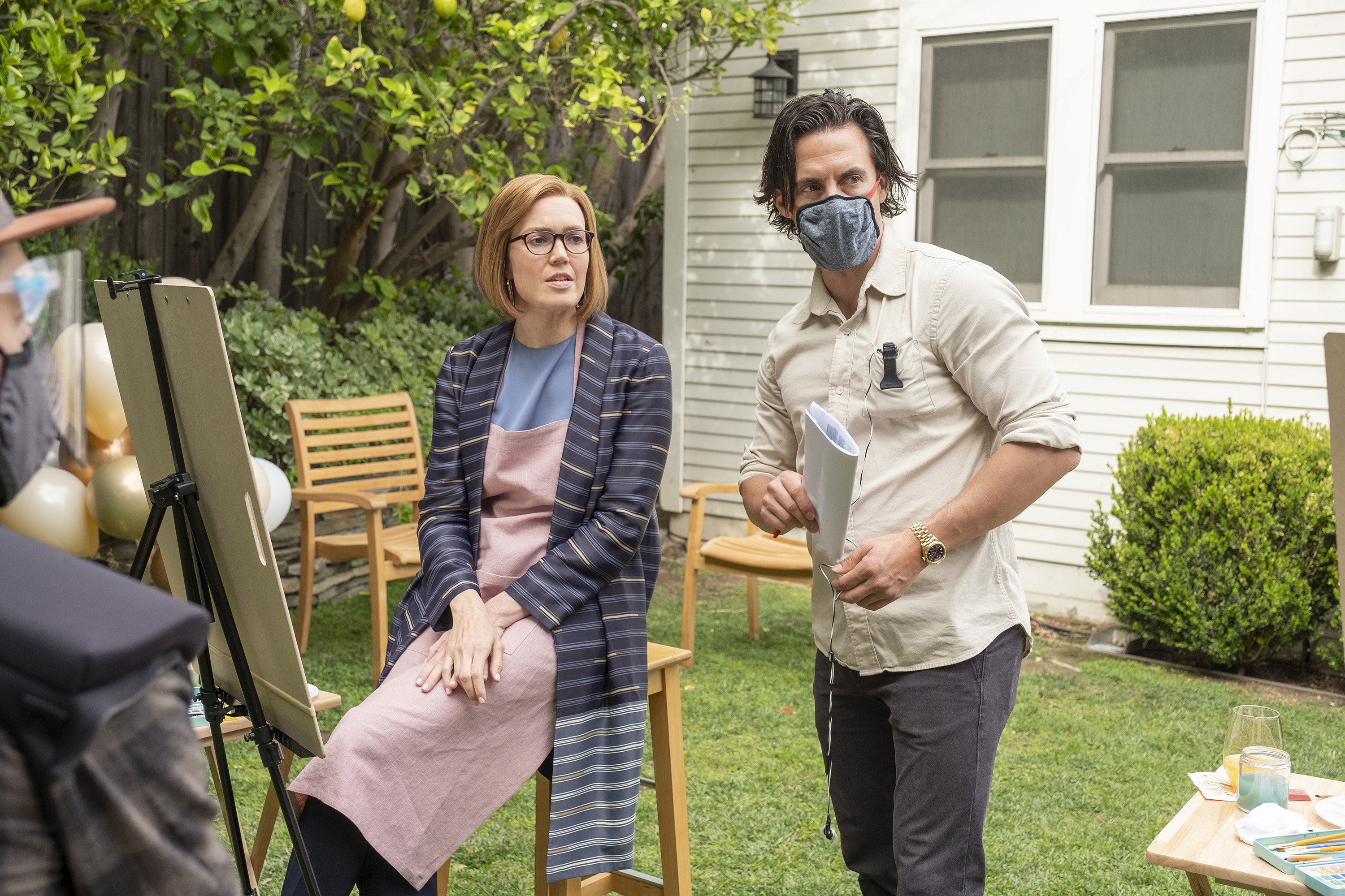 'This Is Us' star Milo Ventimiglia directs his co-star Mandy Moore during a scene. Moore, in character as present day Rebecca, wears a blue striped cardigan over a blue shirt and light pink apron. Ventimiglia wears a white button-up shirt, gray pants, and a gray mask.
