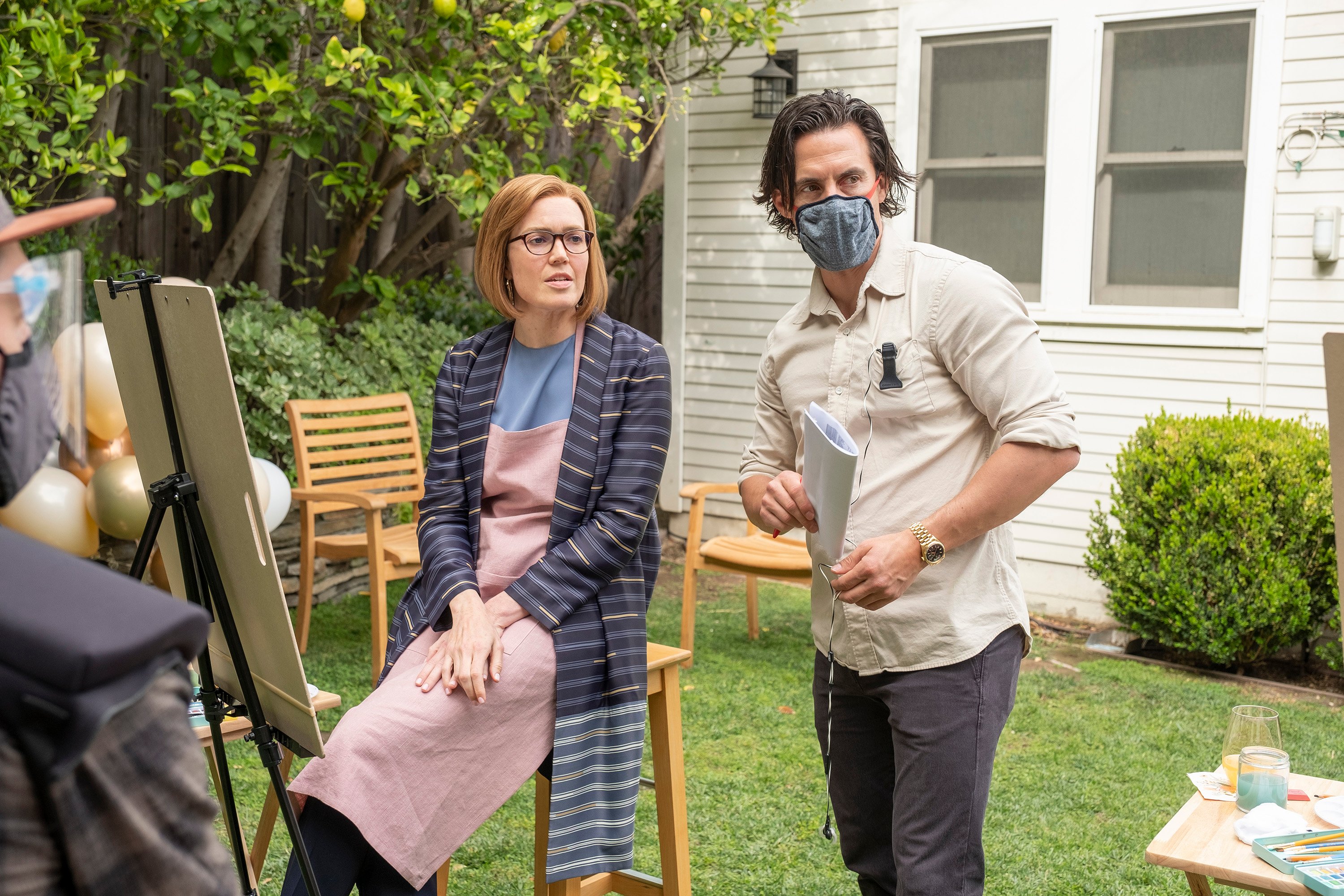 'This Is Us' star Milo Ventimiglia directs his co-star Mandy Moore during a scene. Moore, in character as present day Rebecca, wears a blue striped cardigan over a blue shirt and light pink apron. Ventimiglia wears a white button-up shirt, gray pants, and a gray mask.