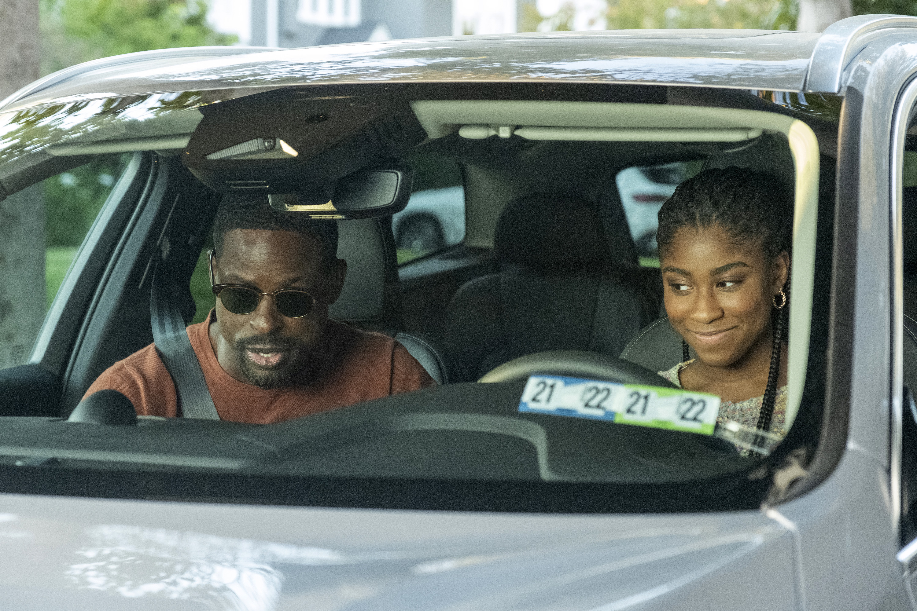 'This Is Us' Season 6 stars Sterling K. Brown and Lyric Ross, in character as Randall and Deja, sit in a car. Randall, in the passenger seat, wears a red sweater and sunglasses. Deja, in the driver seat, wears a multicolored sweater.