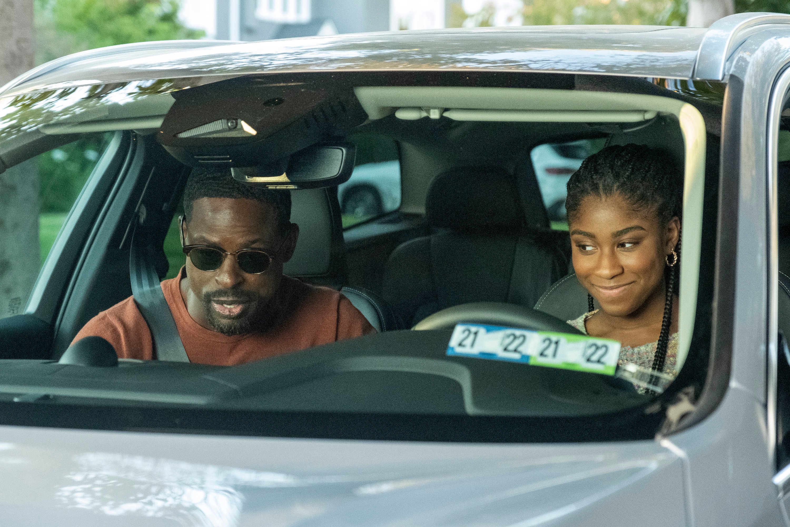 'This Is Us' Season 6 stars Sterling K. Brown and Lyric Ross, in character as Randall and Deja, sit in a car. Randall, in the passenger seat, wears a red sweater and sunglasses. Deja, in the driver seat, wears a multicolored sweater.