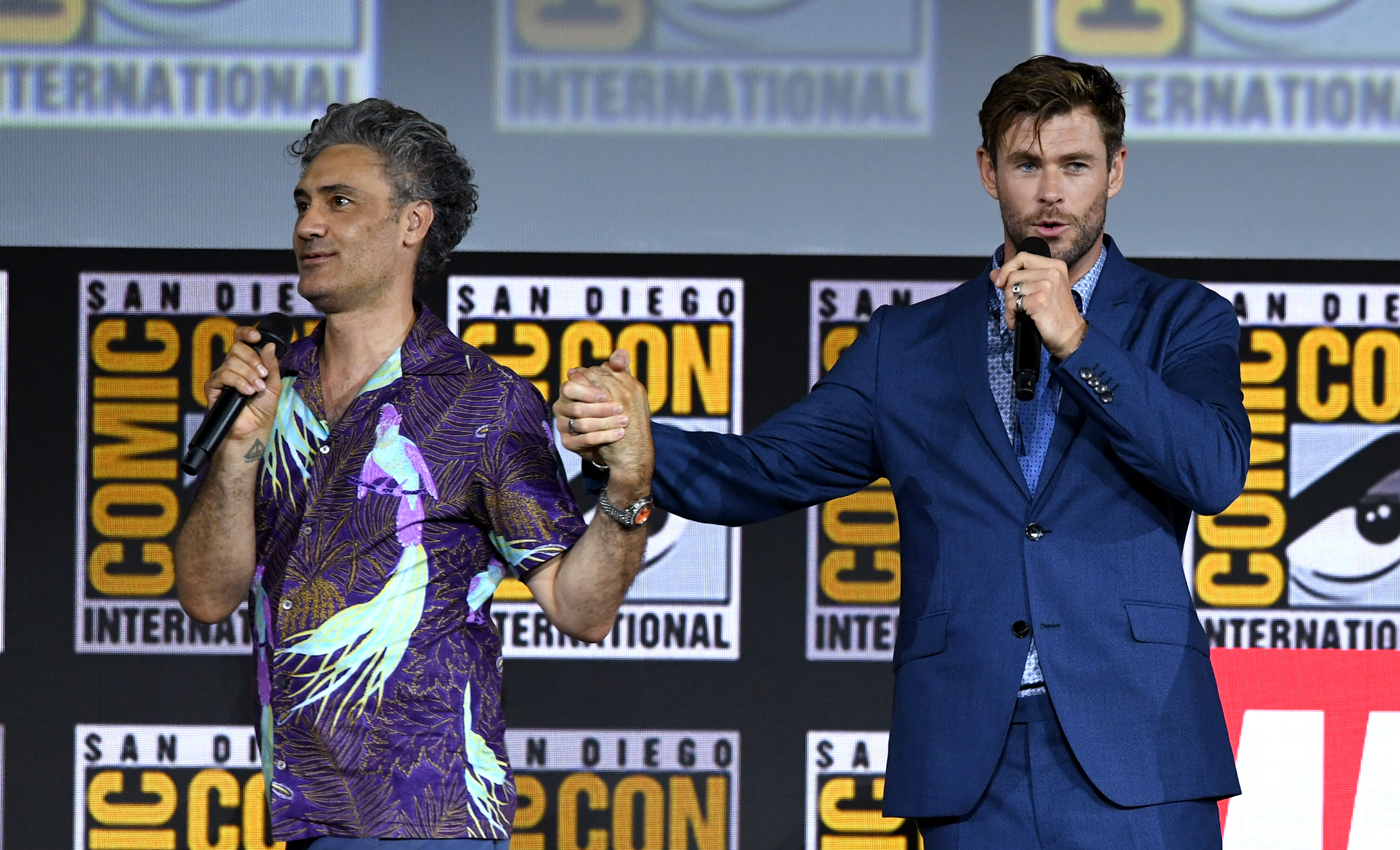 Taika Waititi and Chris Hemsworth, whose movie, Marvel's 'Thor: Love and Thunder,' fans hope to see a trailer for during the 2022 Super Bowl, hold hands while on stage at Comic-Con. Waititi wears a purple and green patterned shirt with birds. Hemsworth wears a blue suit.