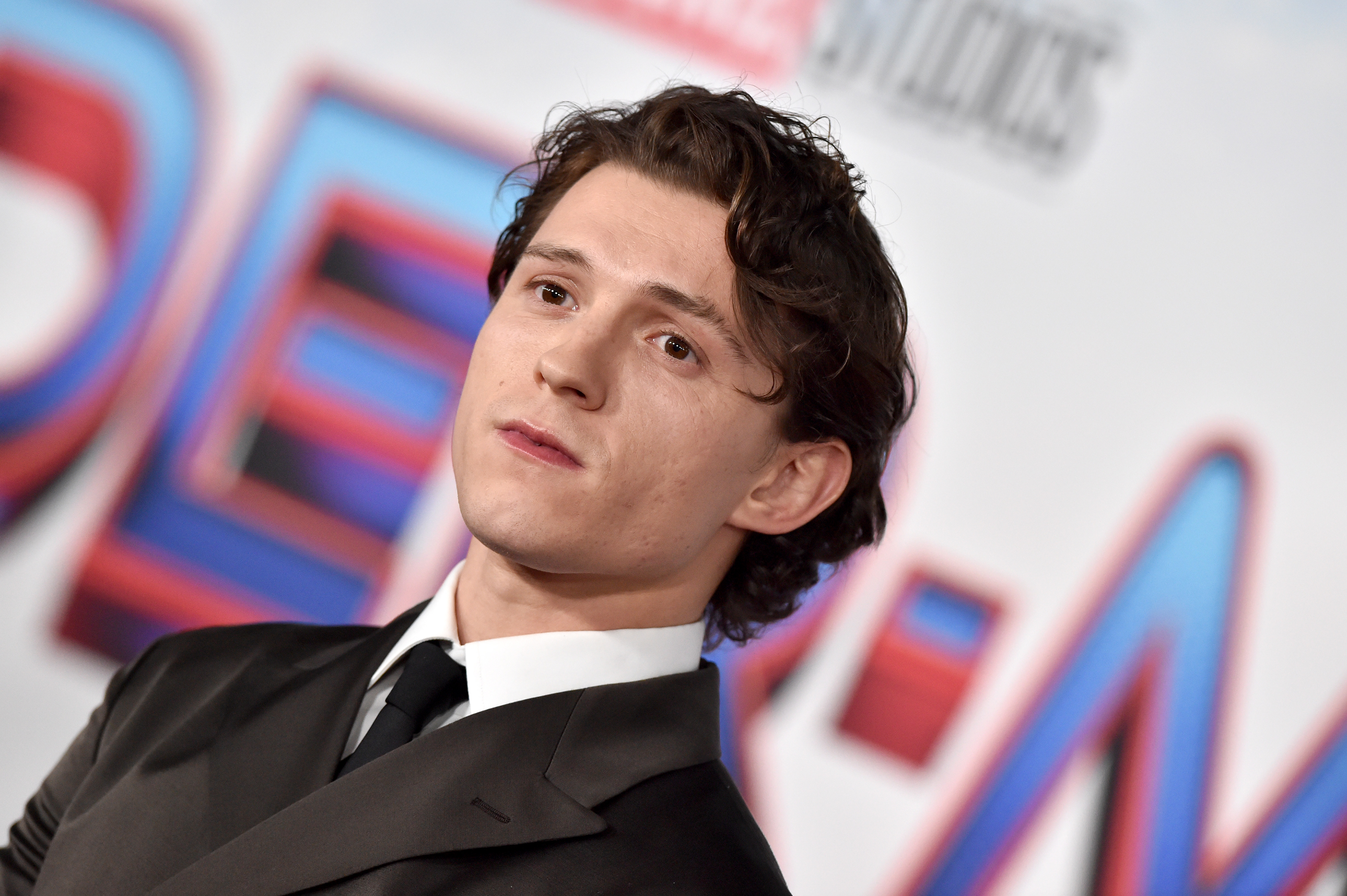 Tom Holland, who is seen in behind-the-scenes content on the 'Spider-Man: No Way Home' Blu-ray, wears a dark gray suit over a white button-up shirt and black tie.