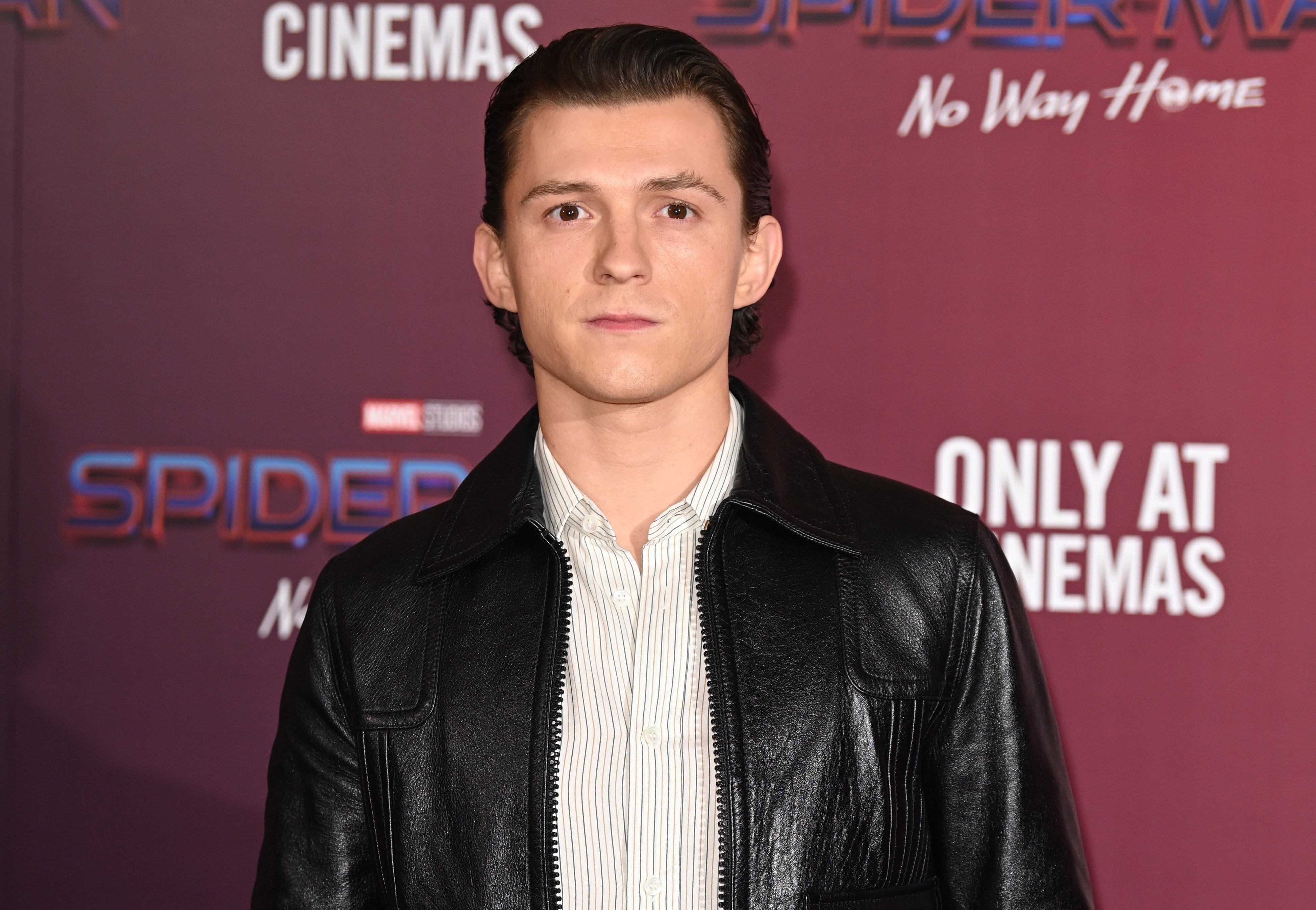 'Spider-Man: No Way Home' star Tom Holland wears a black leather jacket over a white striped button-up shirt.