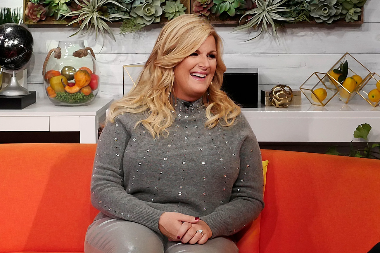 Trisha Yearwood smiles wearing a gray outfit sitting on an orange couch