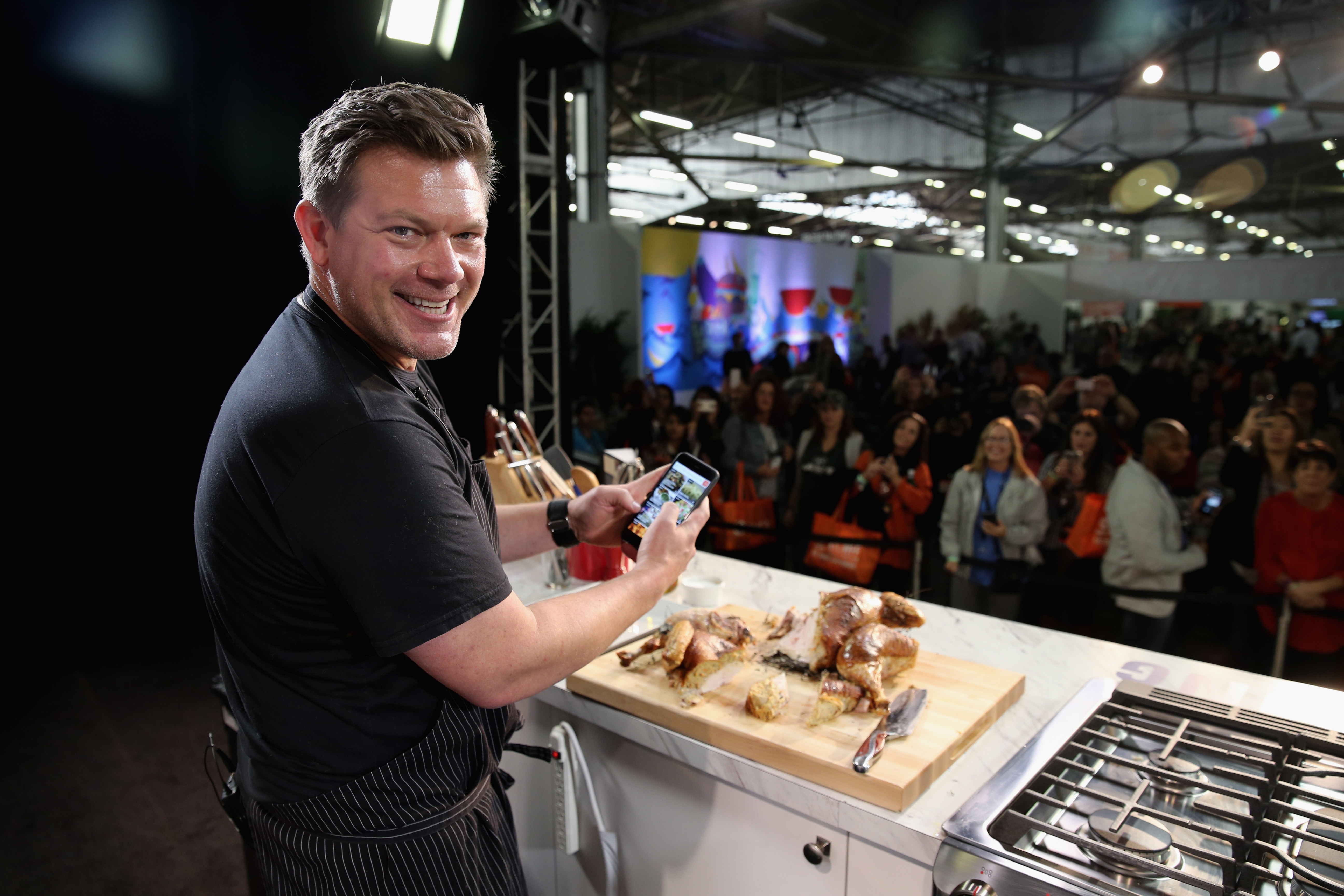 Chef Tyler Florence wears a short-sleeved black shirt as he prepares a meal for a 2015 Food Network event audience.