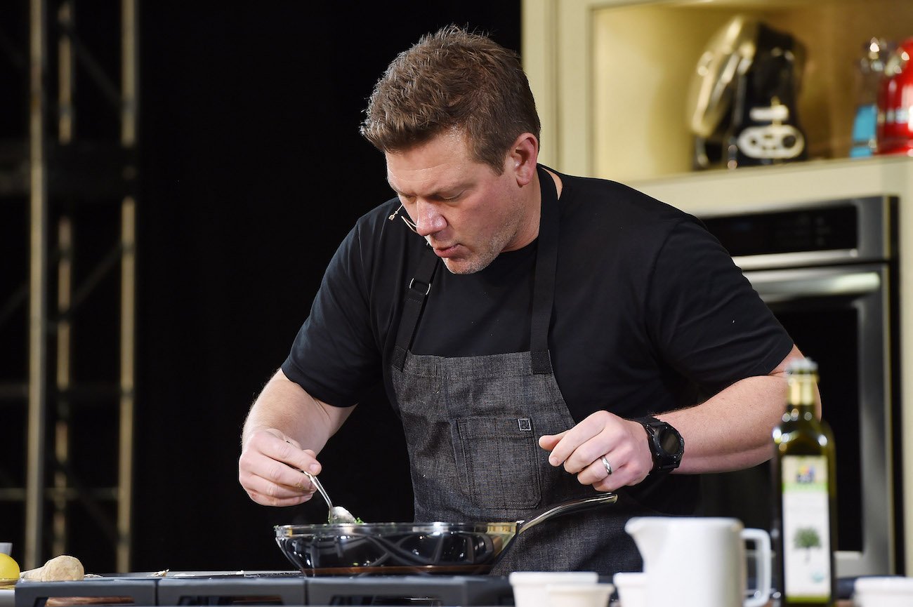 Tyler Florence wears a black shirt and gray apron as he stirs a saute pan