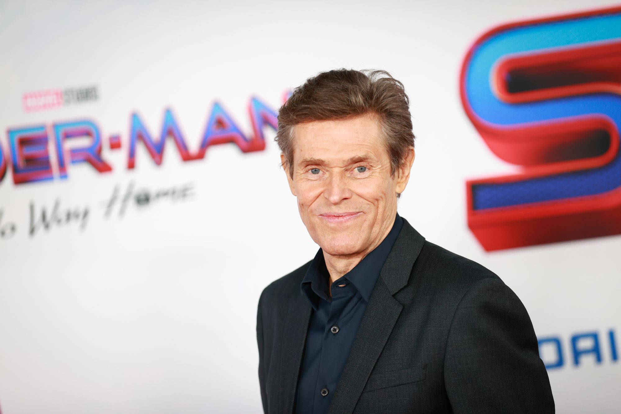 Willem Dafoe, who plays a member of the Sinister Six in 'Spider-Man: No Way Home,' wears a black suit over a dark blue button-up shirt.