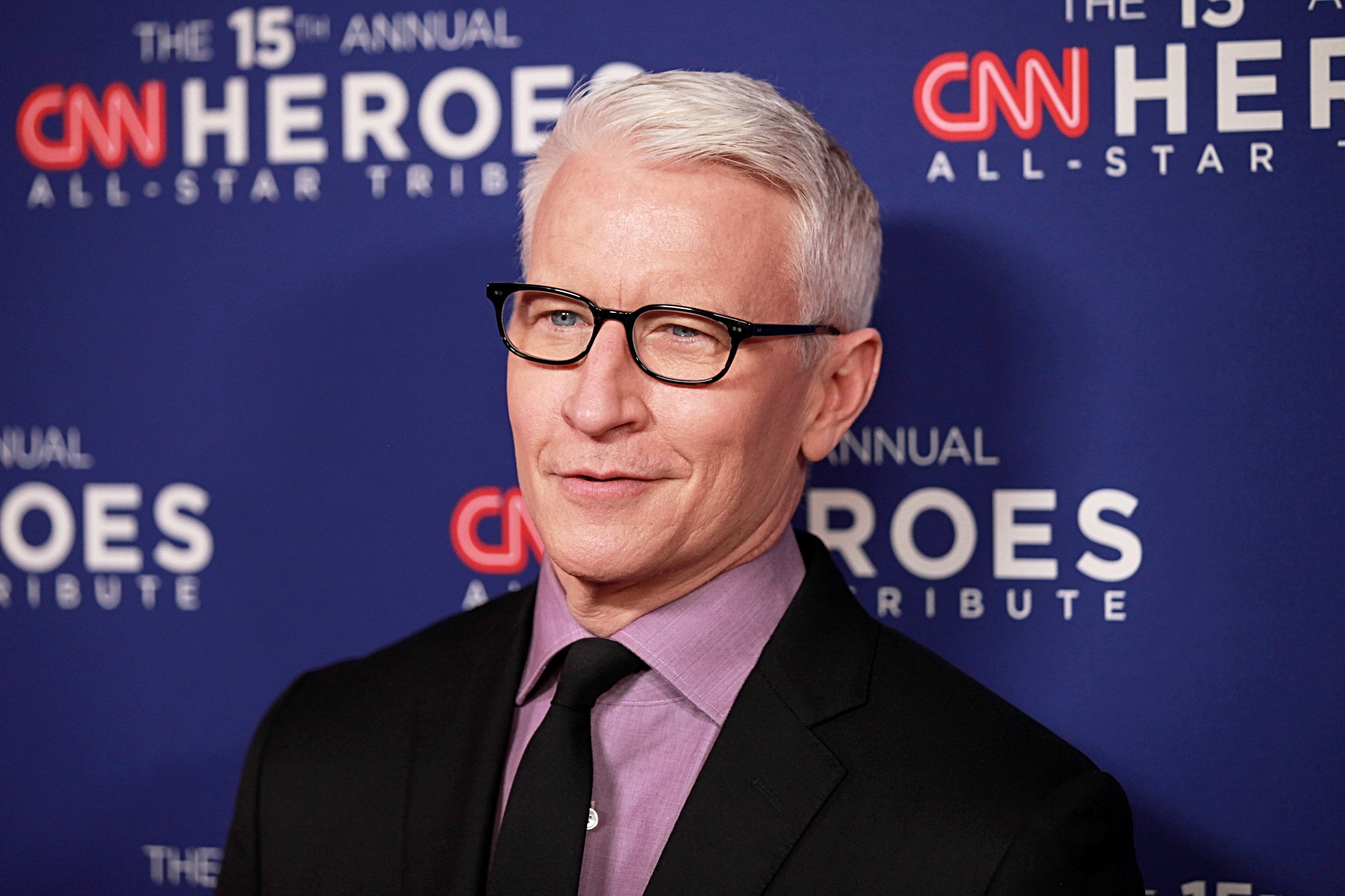 Anderson Cooper posing for the cameras at an event, wearing black frames, a black suit and tie, and a purple shirt