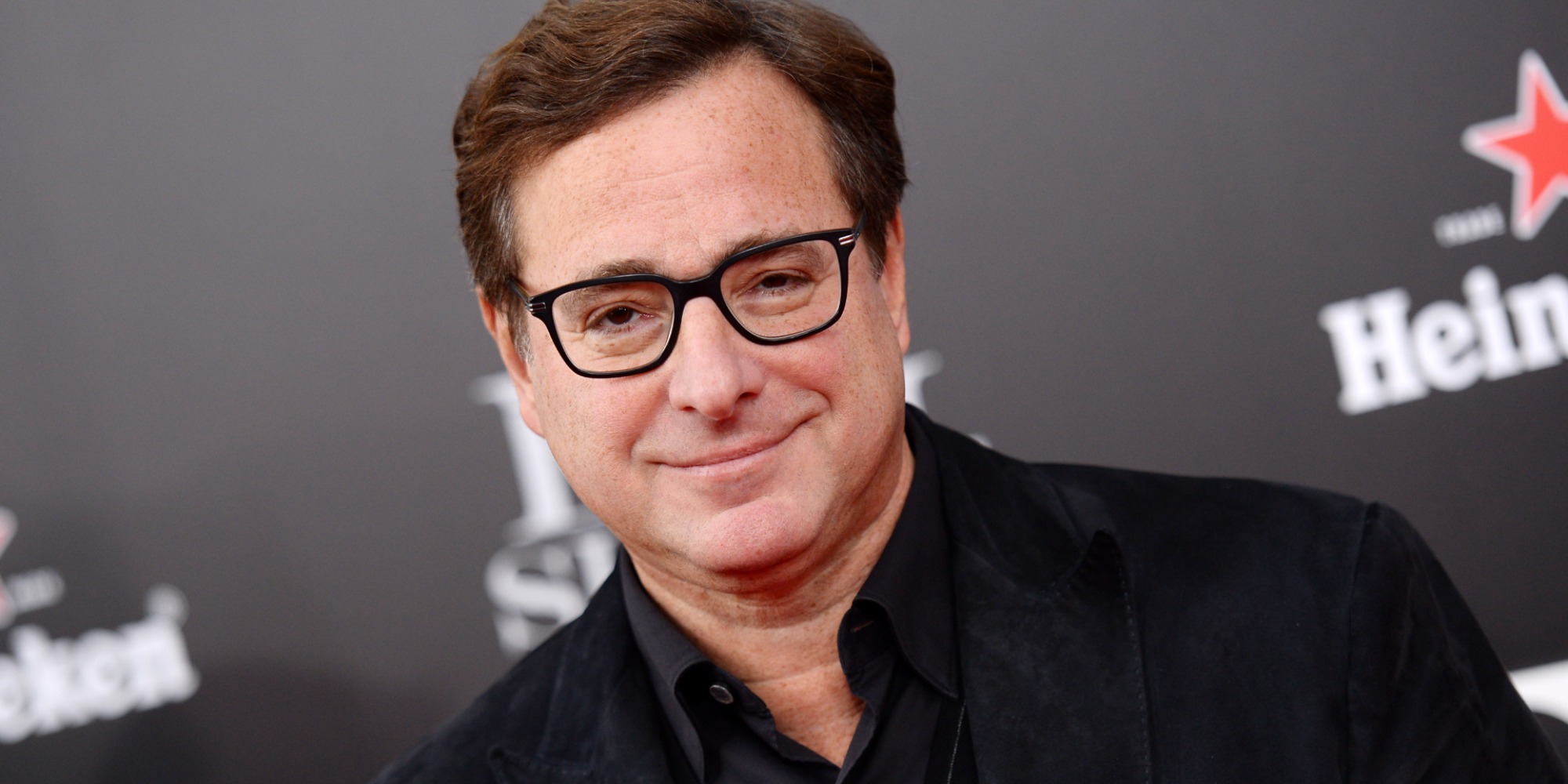 Bob Saget wearing a black suit poses for photographers in front of a black backdrop.