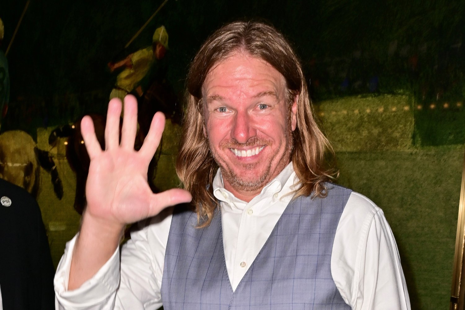 Chip Gaines with long hair, smiling and holding up his hand as they take a photo