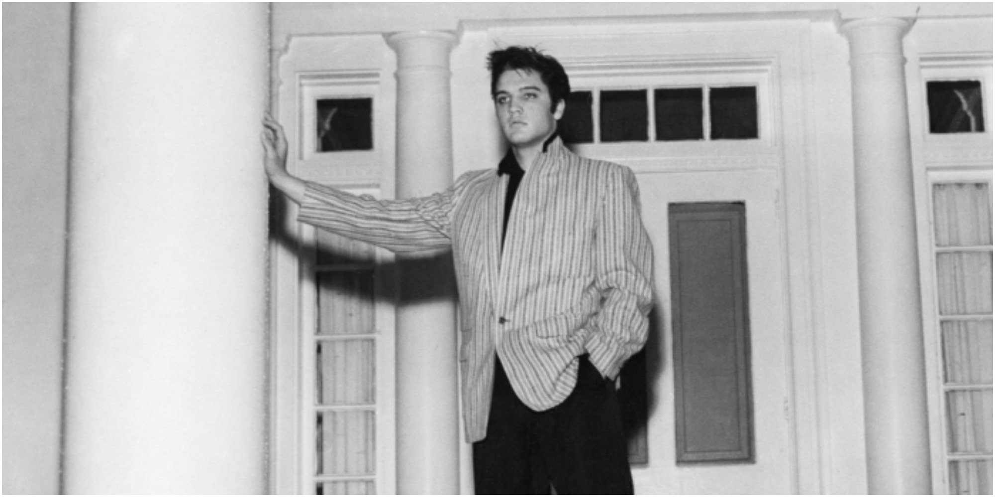Elvis Presley poses in front of Graceland in a black and white image.