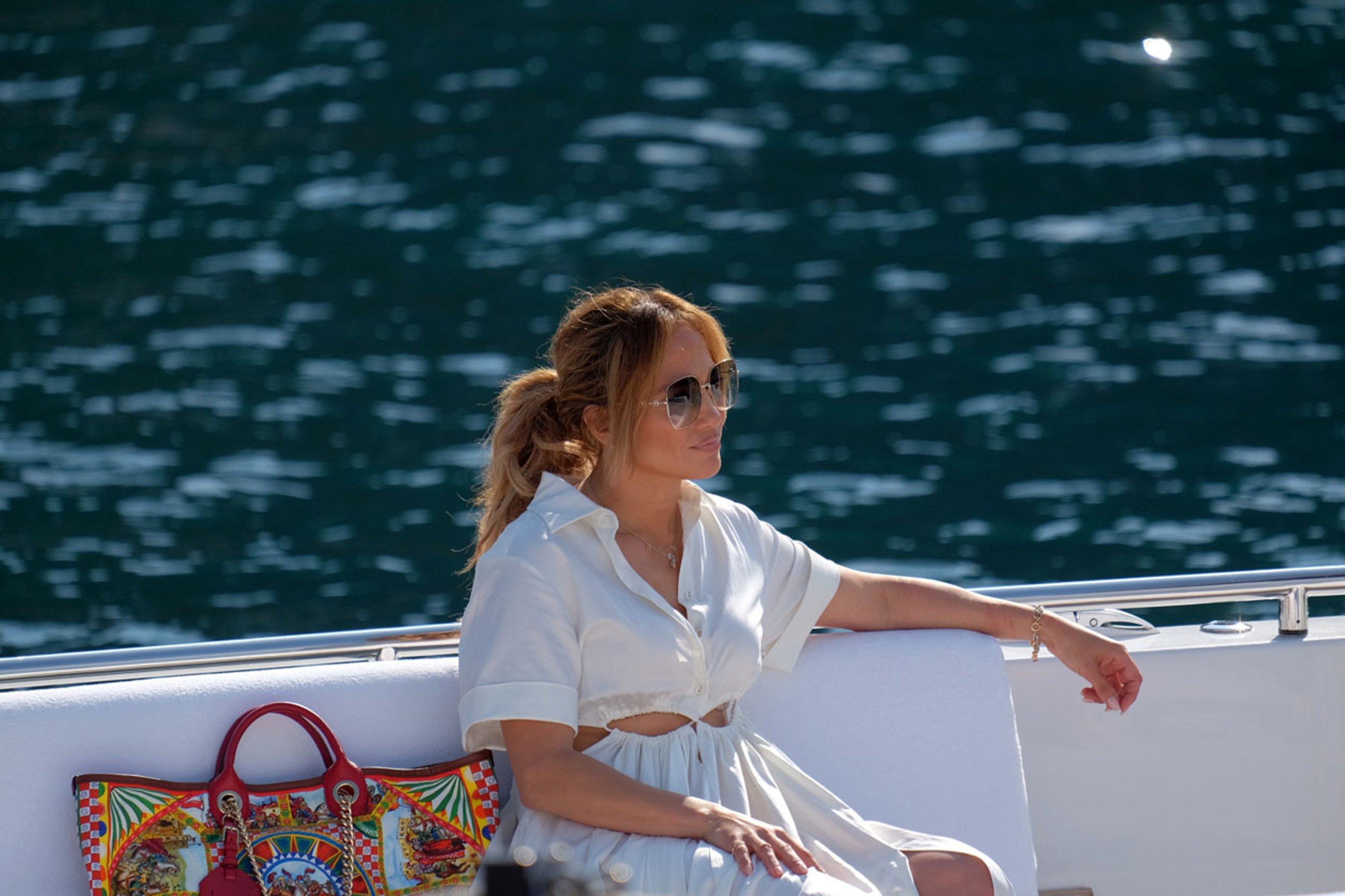 Jennifer Lopez sits in a boat while wearing a white dress and sunglasses. 