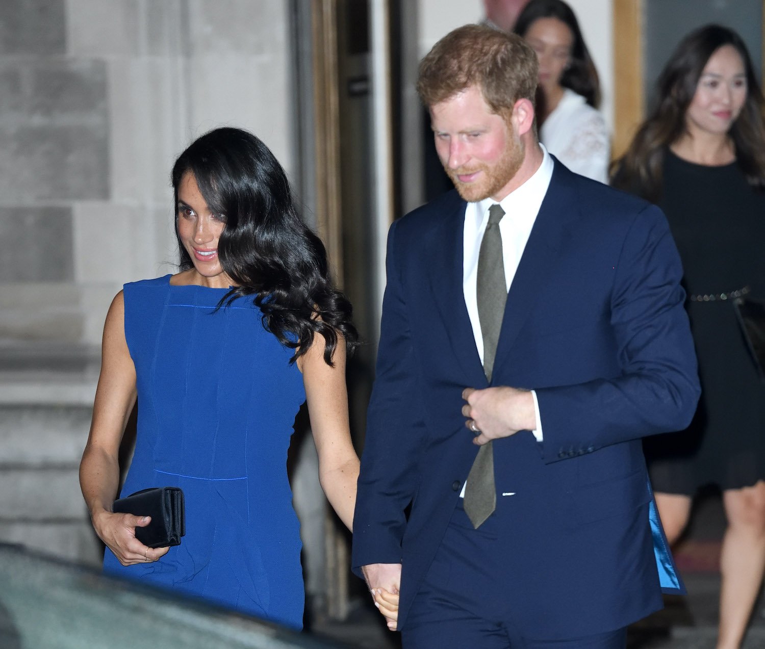 Meghan Markle wears a blue dress and looks to the side while smiling as she holds hands with Prince Harry, wearing a suit and tie, after leaving the 100 Days to Peace concert in 2018