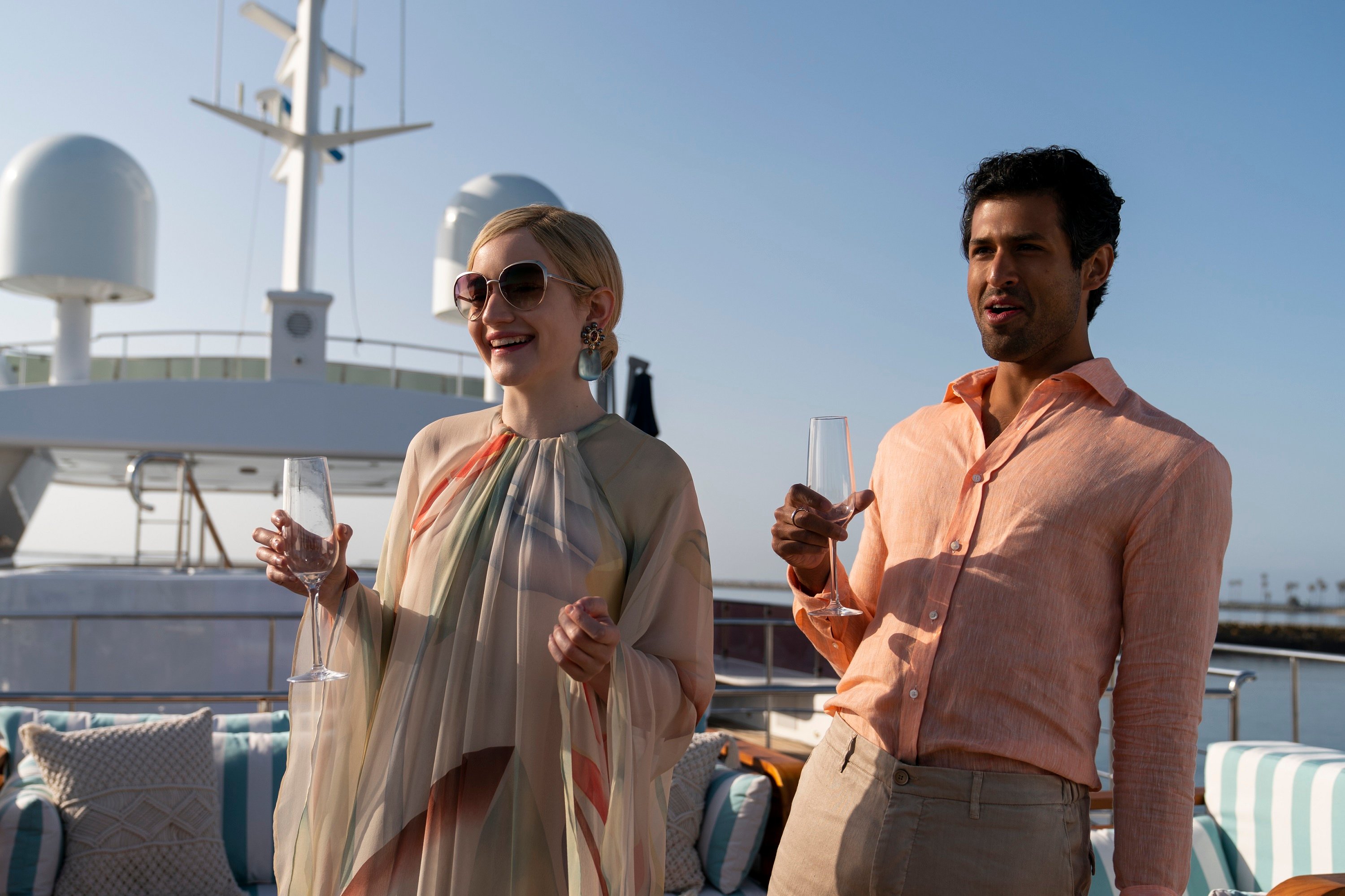 'Inventing Anna' cast members Julia Garner and Saamer Usmani holding drinks on a yacht as Anna Delvey and Chase Sikorski