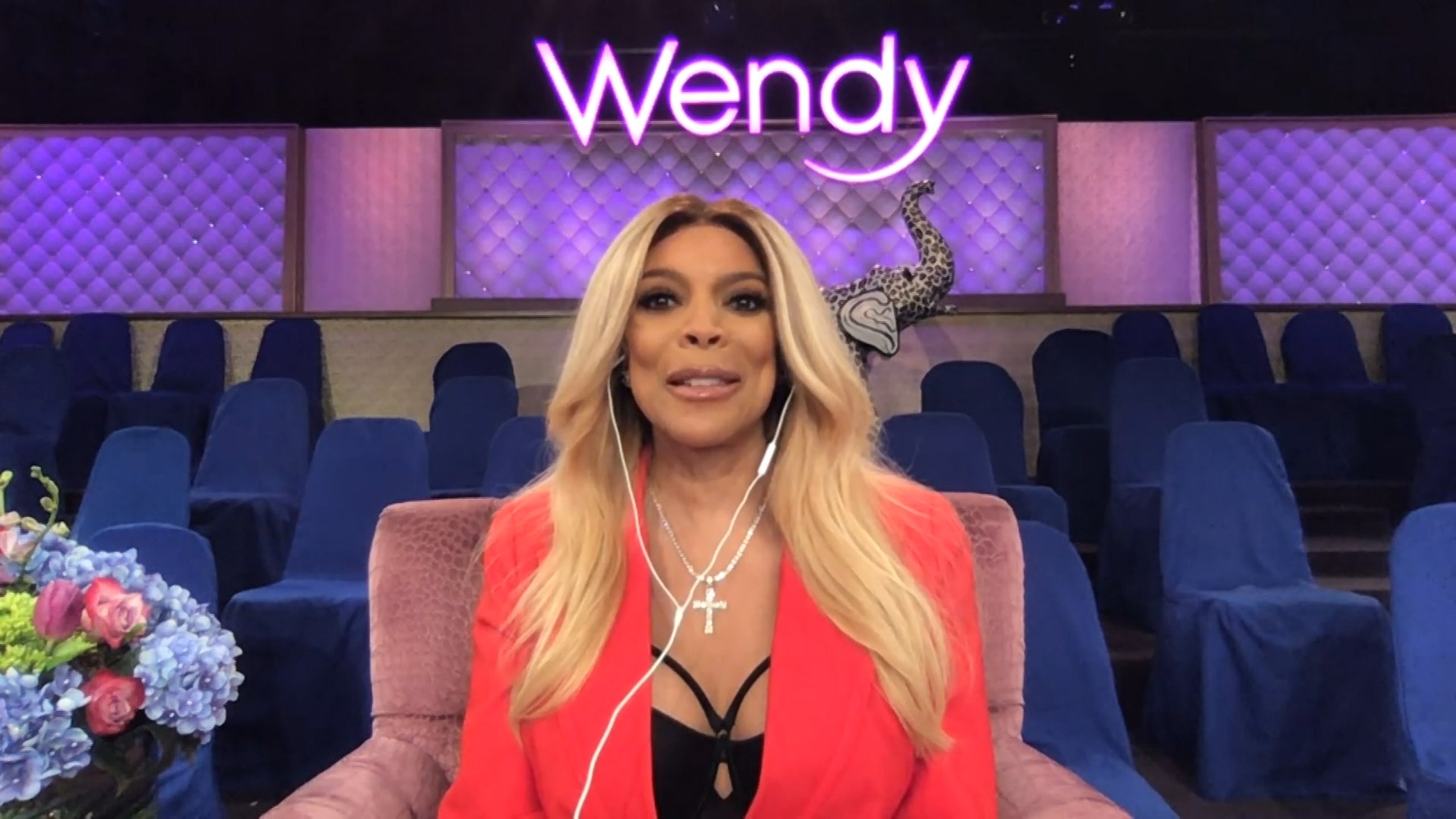 Wendy Williams sitting in her purple chair on the set of her show, wearing an orange blazer and black top