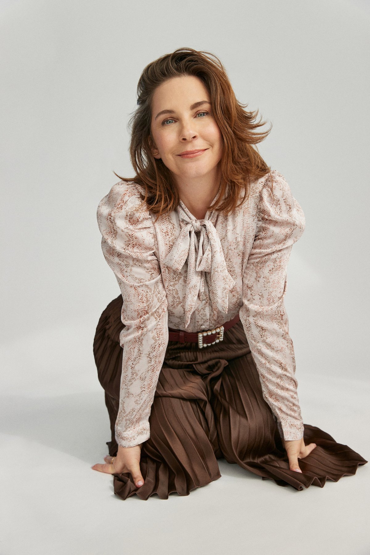 Portrait of Allison Gabriel from 'Sweet Magnolias' kneeling and wearing a tie-neck blouse and long brown skirt