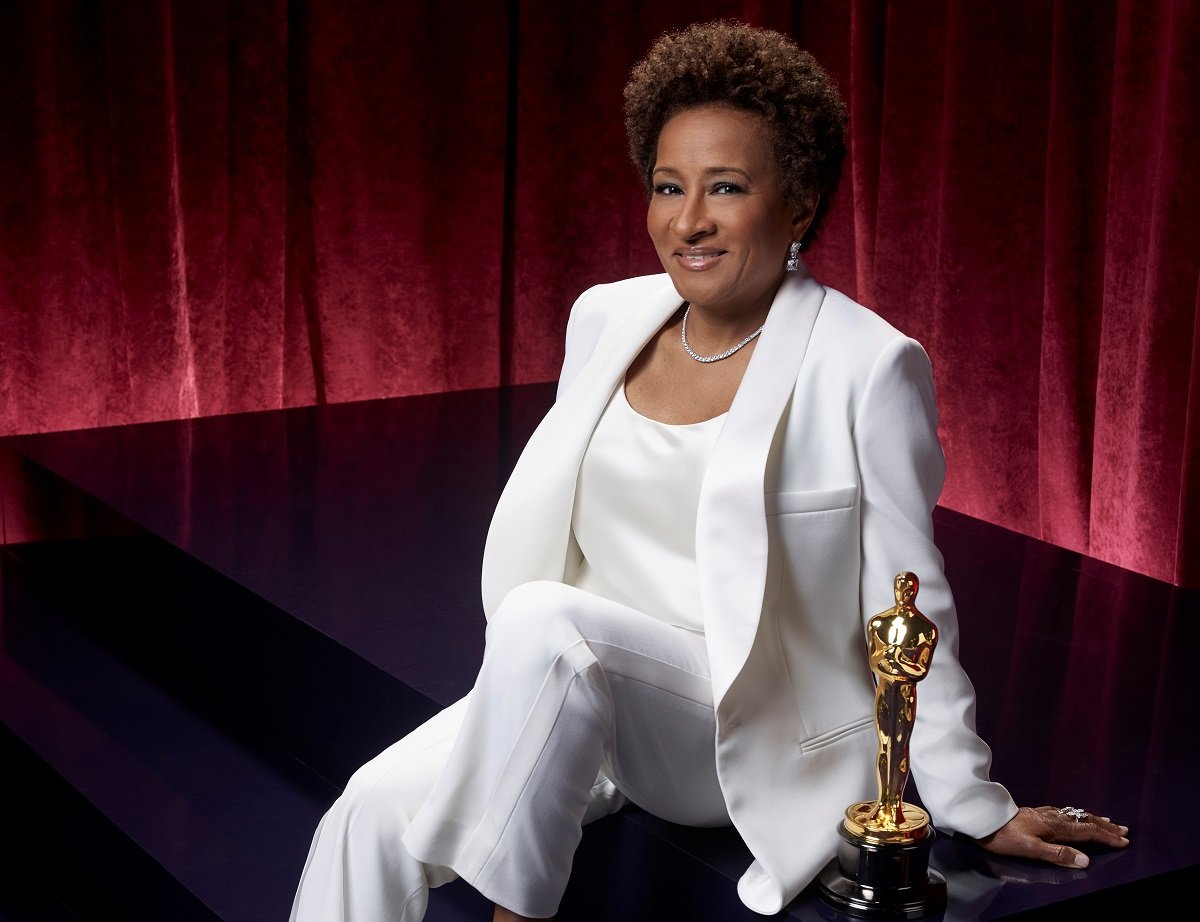 Academy Award host Wanda Sykes, whose net worth has grown thanks to her film and other roles, sitting in a white suit next to an Oscar