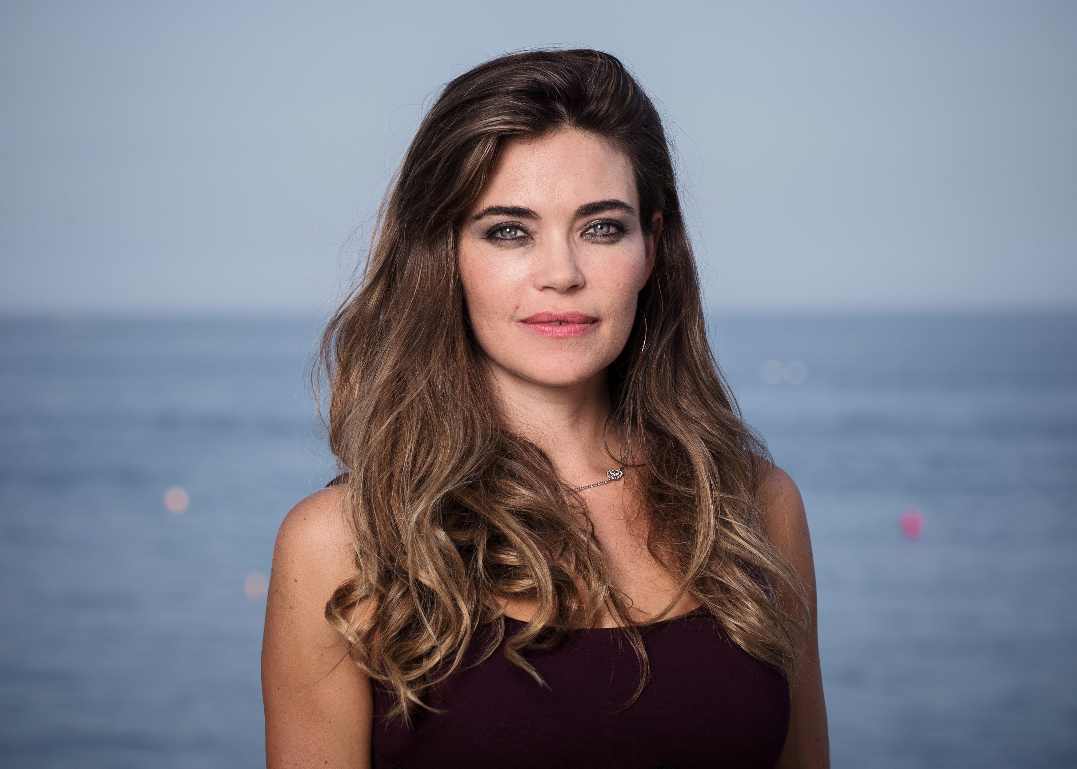 'The Young and the Restless' actor Amelia Heinle wearing a burgundy dress and posing in front of an ocean backdrop.
