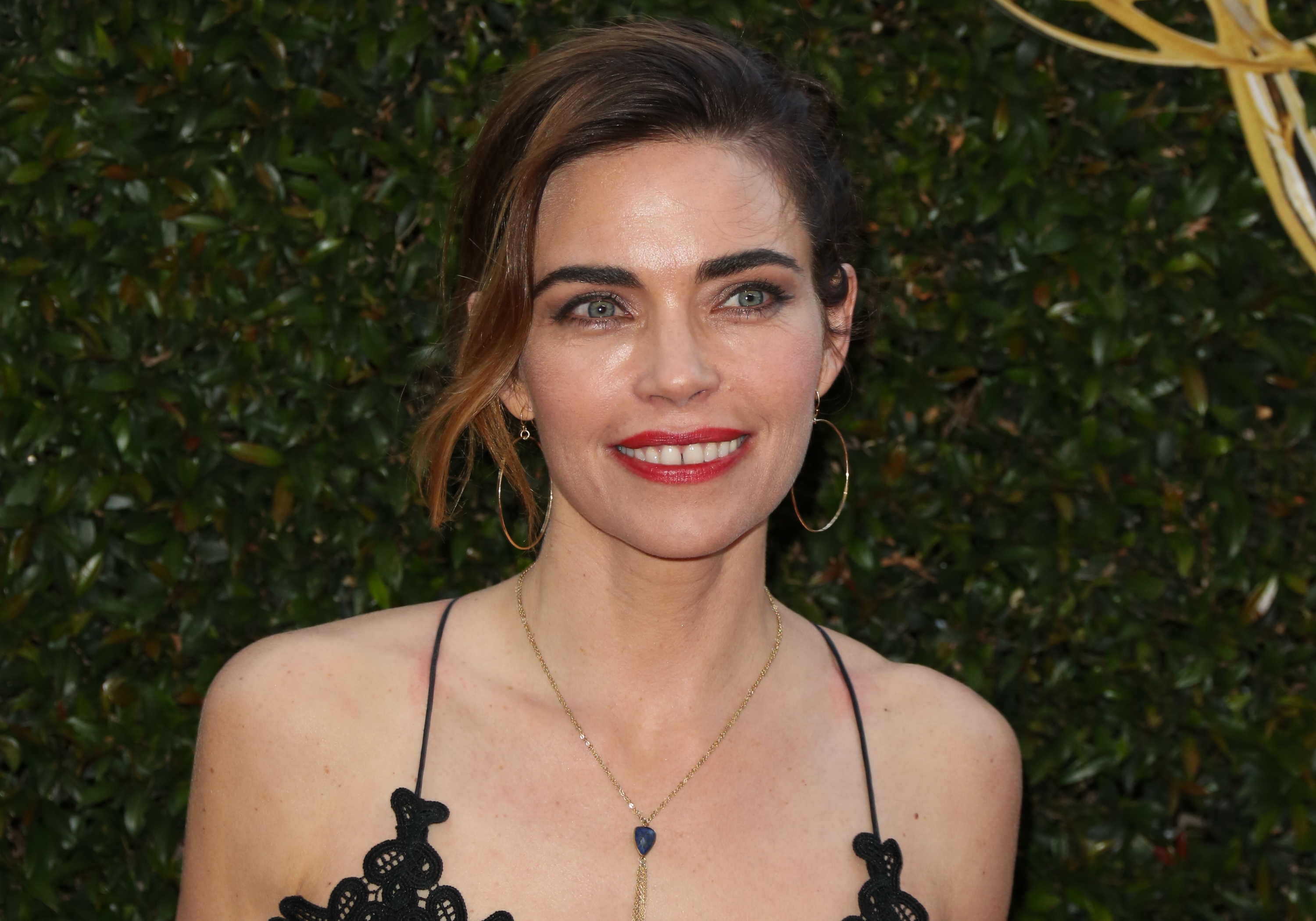 'The Young and the Restless' actor Amelia Heinle wearing a black dress and standing in front of a green hedge.