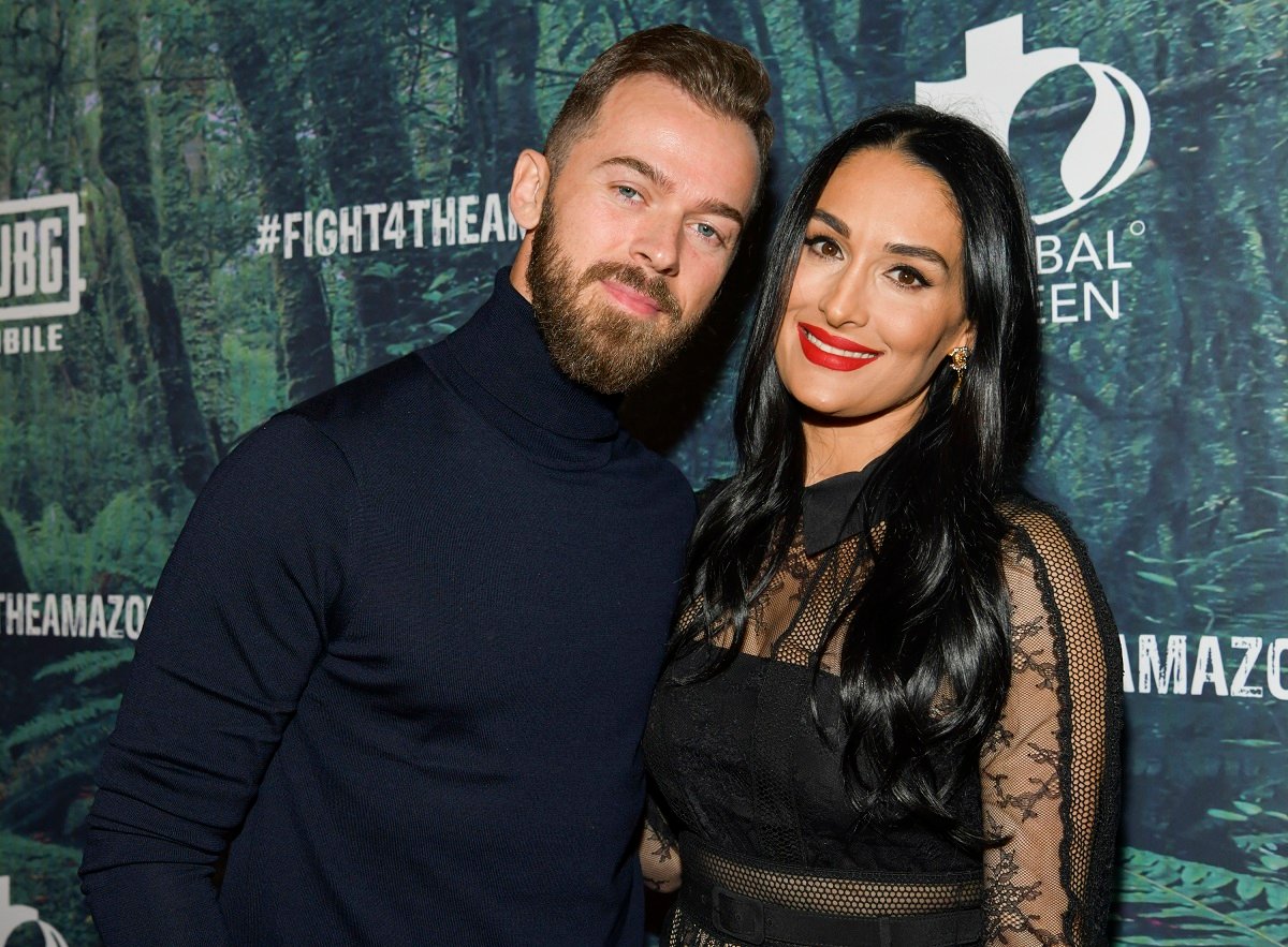 Artem Chigvintsev dressed in a black turtleneck poses with fiancée Nikki Bella, who is also dress in a blac outfit. 
