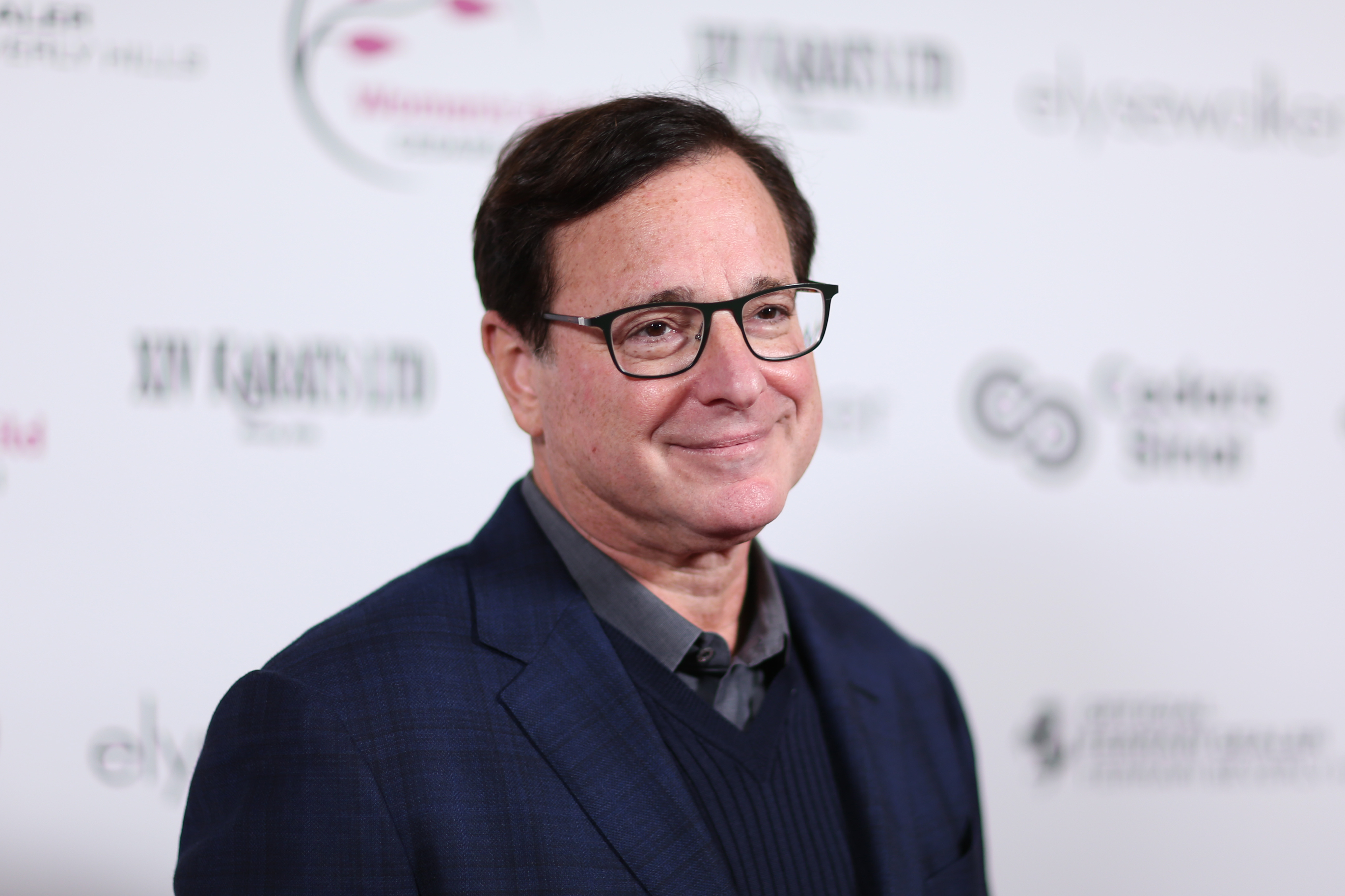 Comedian and 'Full House' star Bob Saget. He's wearing a dark blue suit with a collared shirt and black sweater under it.