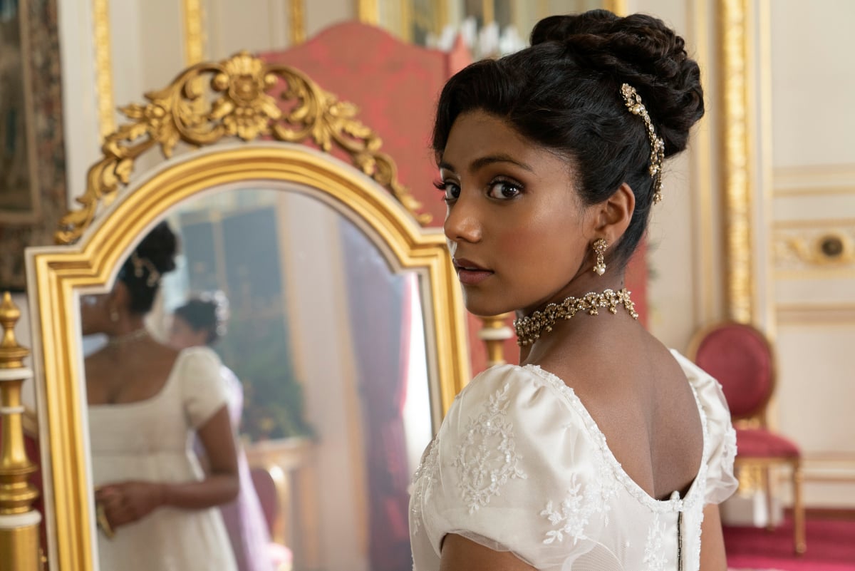 Charithra Chandran as Edwina Sharma in Bridgerton Season 2. Edwina stands in front of a mirror wearing a white dress. 