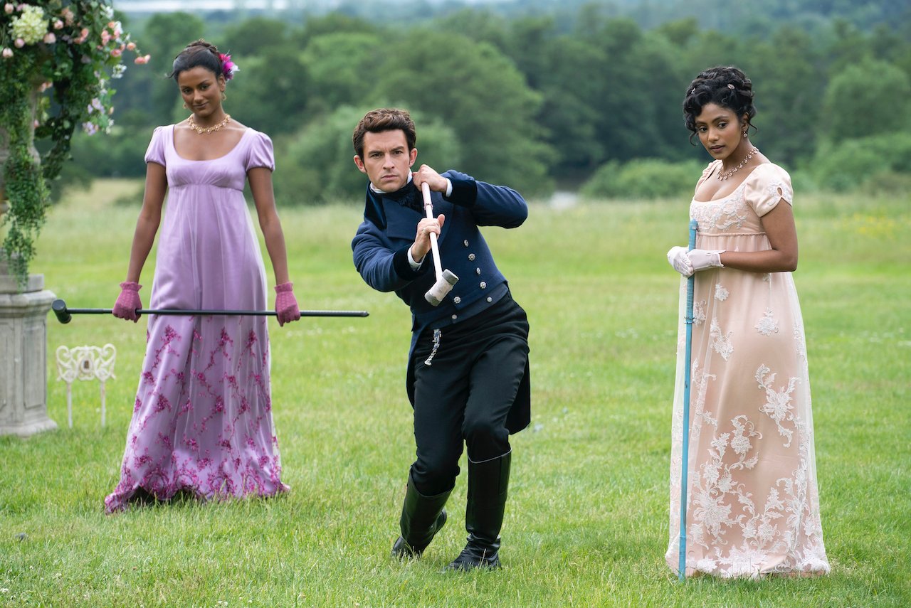 (L to R) Simone Ashley as Kate Sharma, Jonathan Bailey as Anthony Bridgerton, Charithra Chandran as Edwina Sharma play cricket together in a courtyard in 'Bridgerton'.