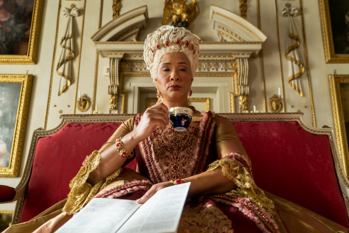 Golda Rosheuvel as Queen Charlotte, wife of King George III, in Bridgerton. Charlotte sits on her throne sipping tea. 