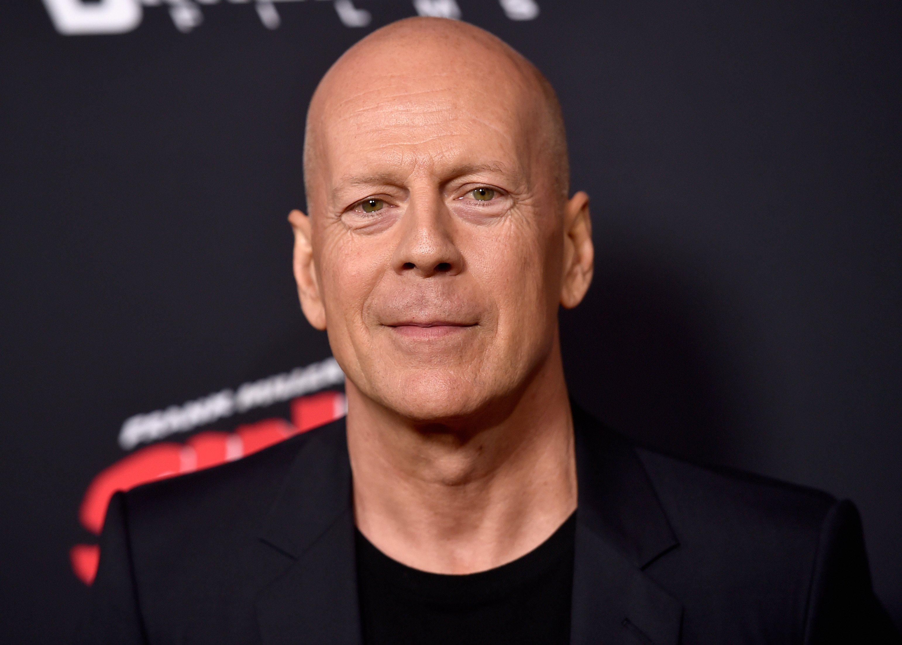 Bruce Willis poses in front of a black background during a media event. 