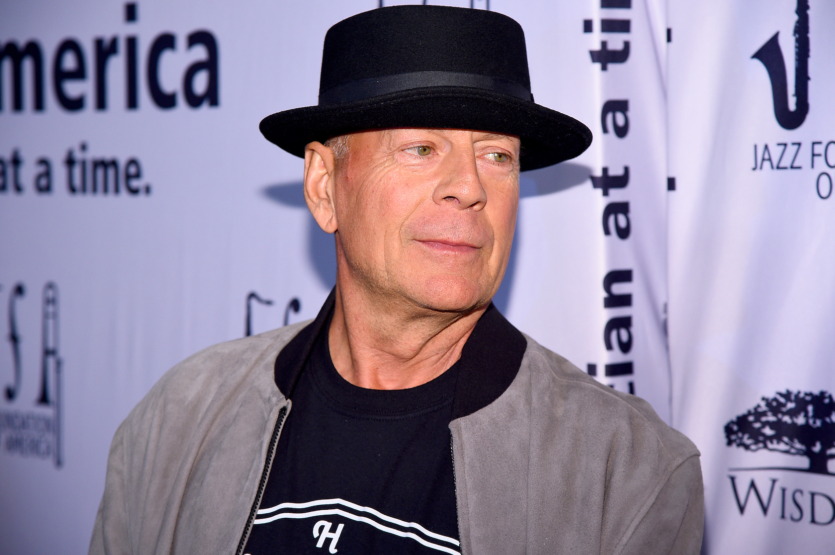 Bruce Willis in a black hat looking over his shoulder against a purple background. Bruce Willis is leaving Hollywood due to aphasia