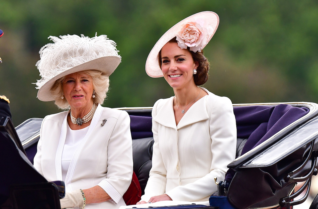 Camilla Parker Bowles and Meghan Markle sitting in a carriage and wearing white outfits with hats