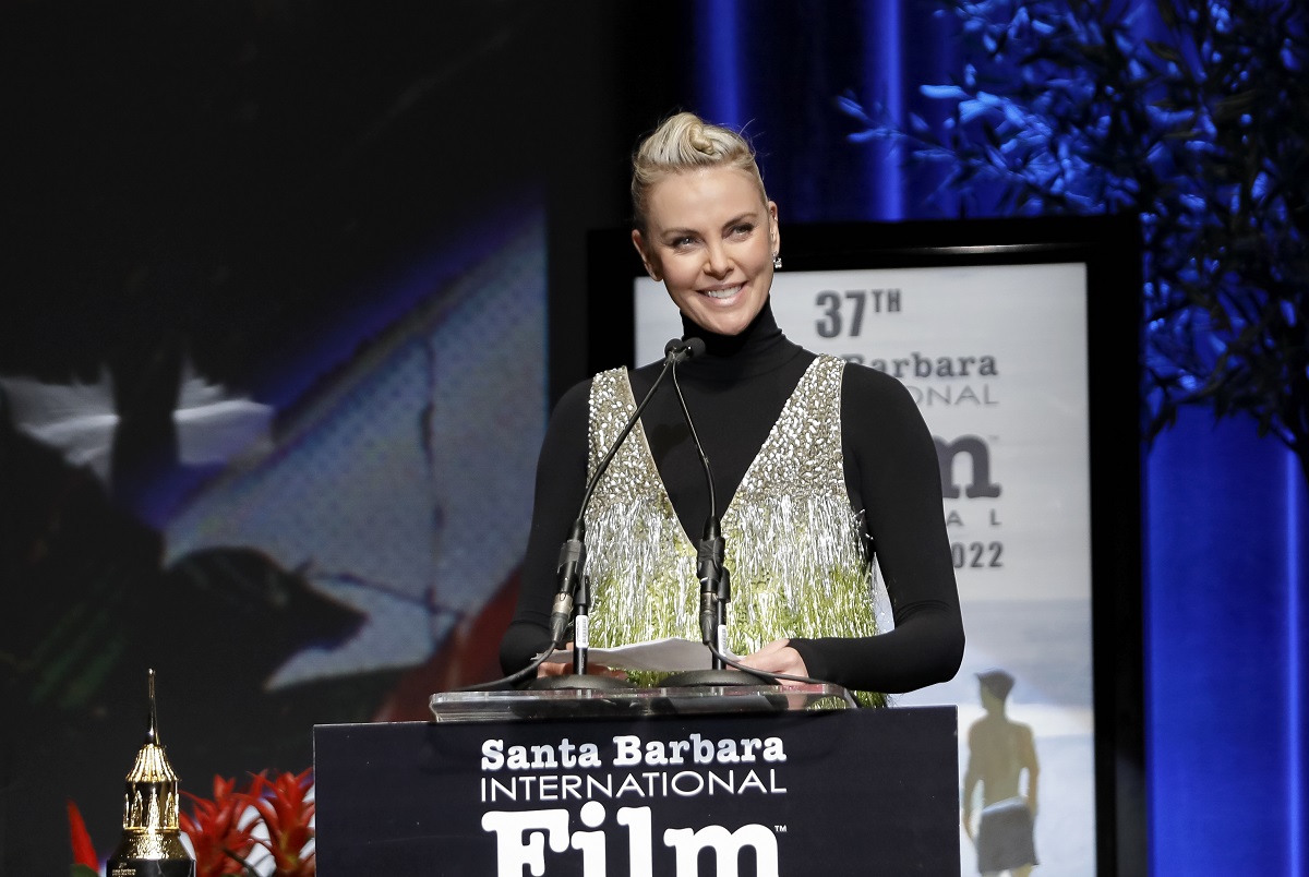 Charlize Theron speaking on stage while wearing a black and gold dress.