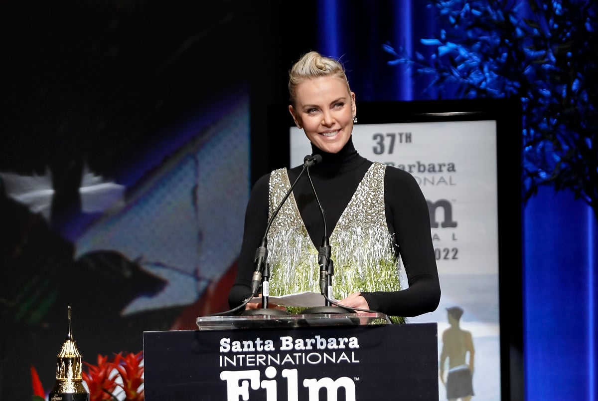 Charlize Theron speaking on stage while wearing a black and gold dress.