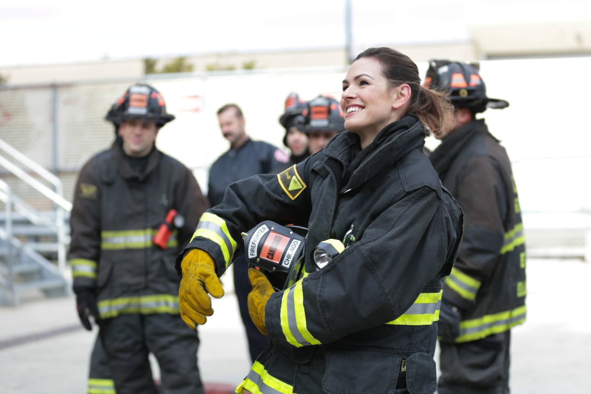 Daisy Betts as Rebecca Jones in Chicago Fire. Jones smiles wearing firefighter gear and holding her helmet under her arm.