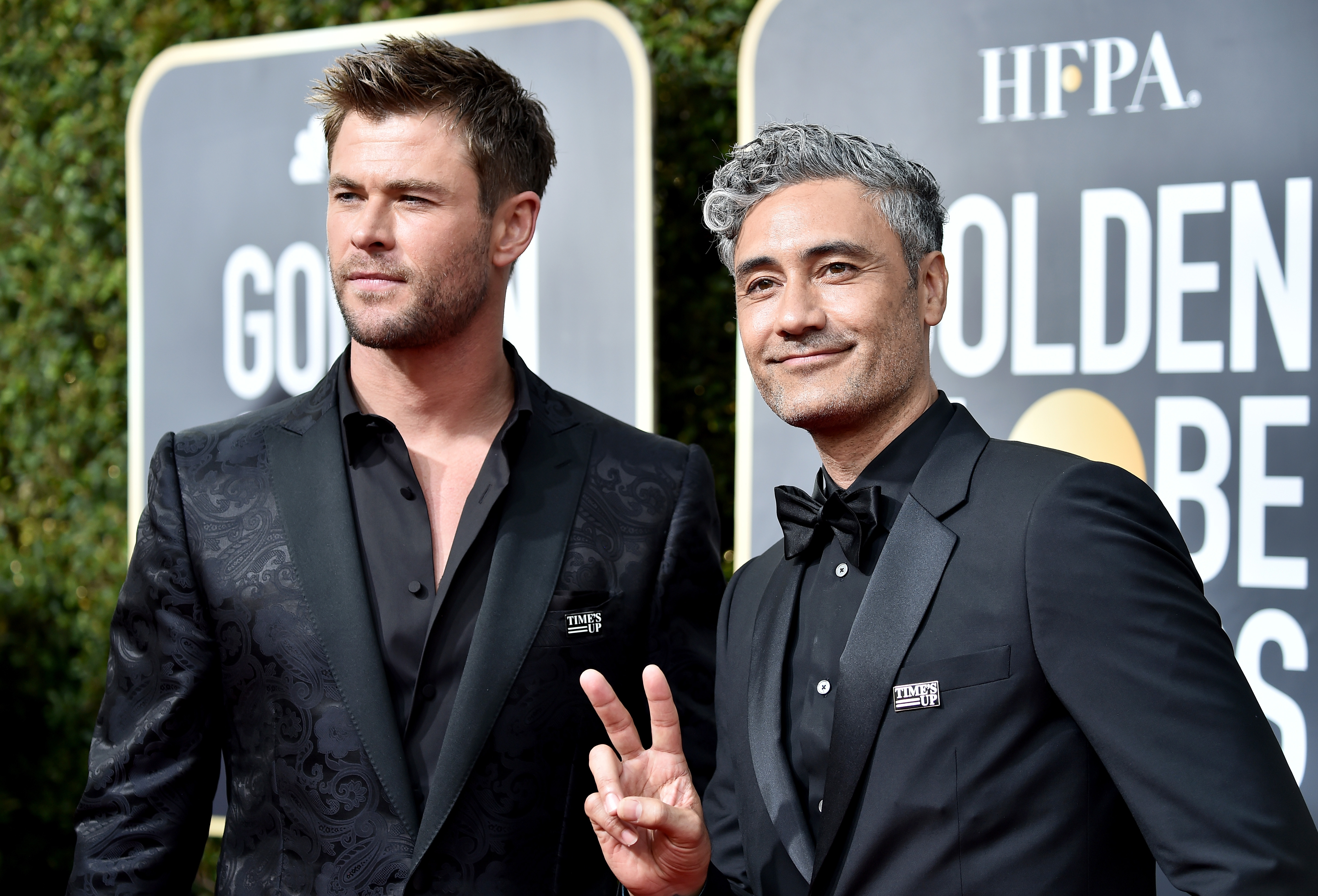 'Thor: Love and Thunder' stars Chris Hemsworth and Taika Waititi pose for pictures together on the red carpet. Hemsworth wears a floral black suit over a black button-up shirt. Waititi wears a black suit over a black button-up shirt and black bow tie.