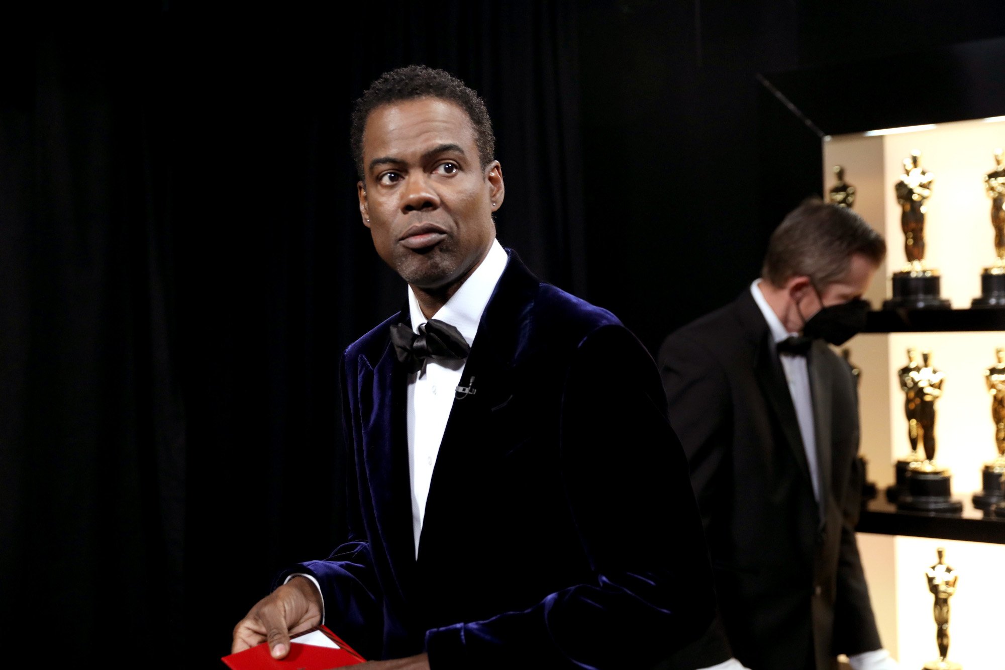 Comedian Chris Rock at the 2022 Oscars. He's wearing a suit, holding a card, and standing backstage.