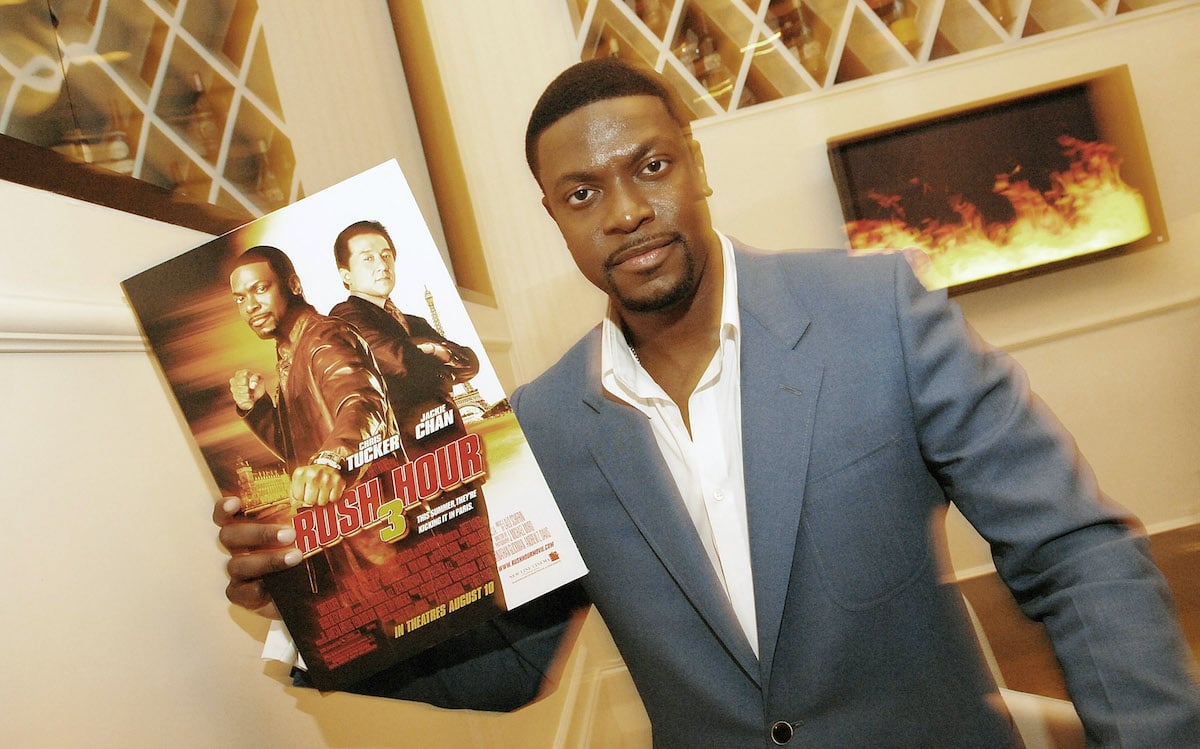 Chris Tucker wears a grey suit and poses with a ‘Rush Hour 3’ poster