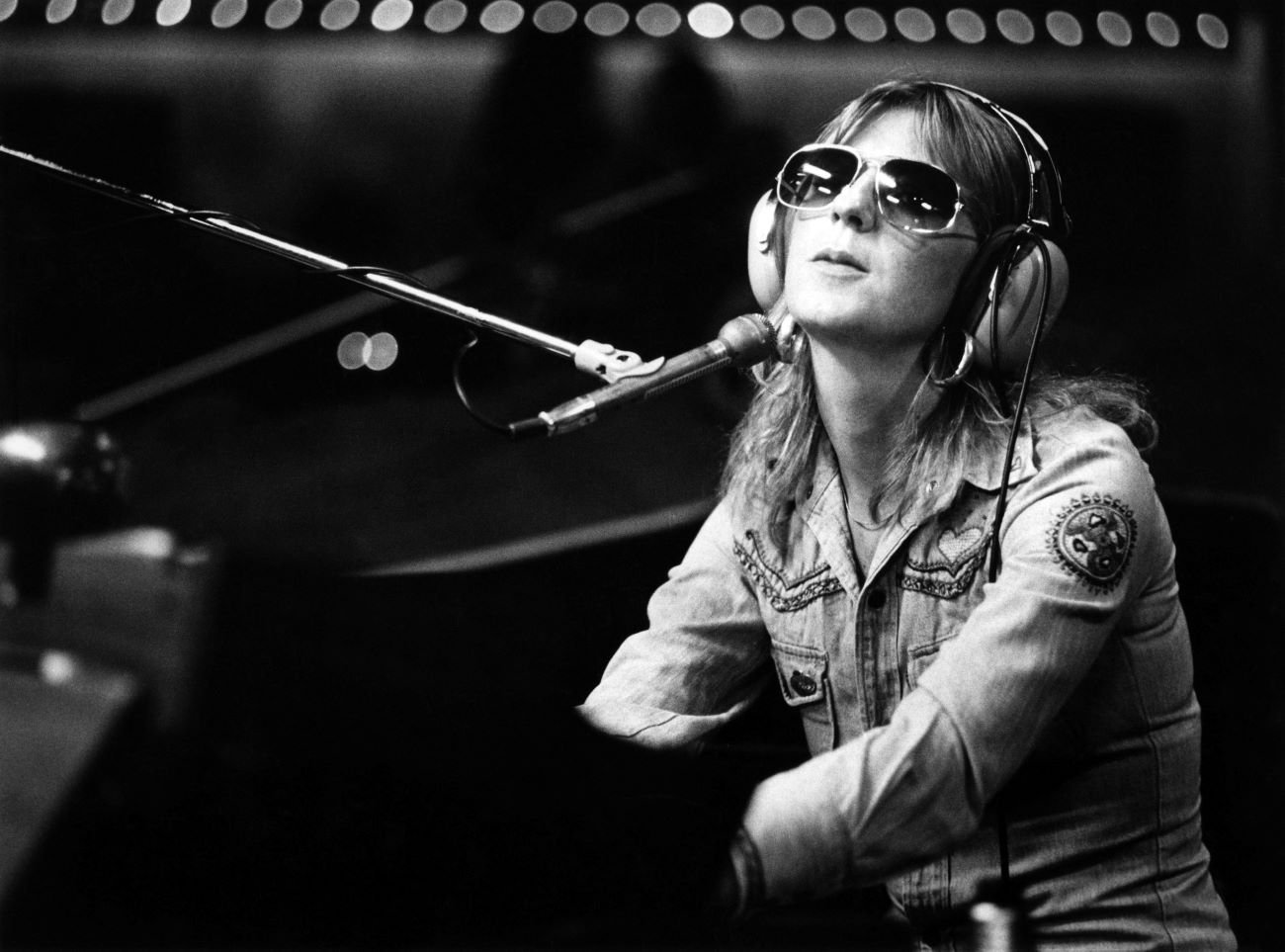 A black and white photo of Fleetwood Mac member Christine McVie wearing a denim jacket, sunglasses, and headphones. She sits in front of a microphone.