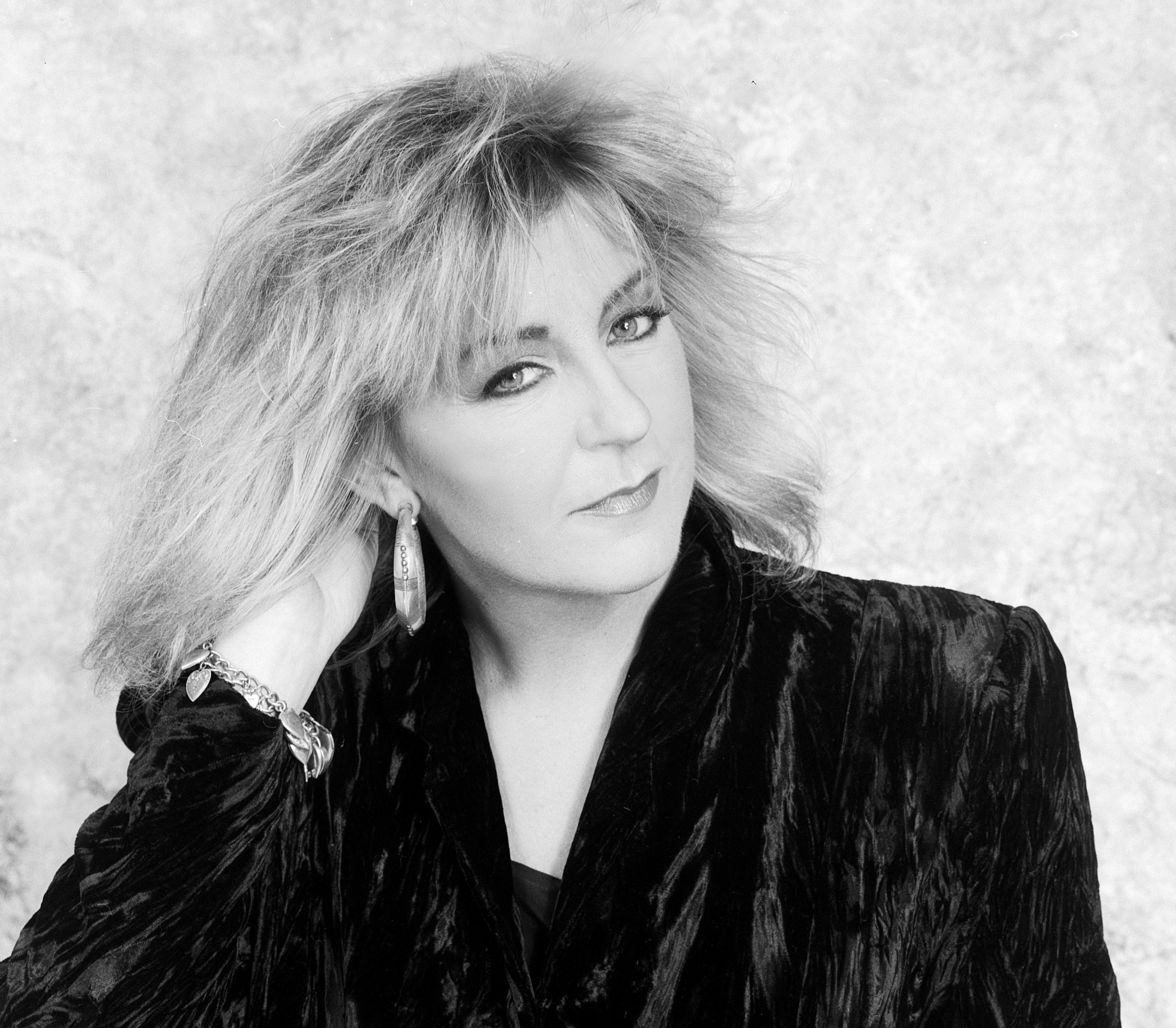A black and white photo of Christine McVie wearing a velvet jacket and hoop earrings. 